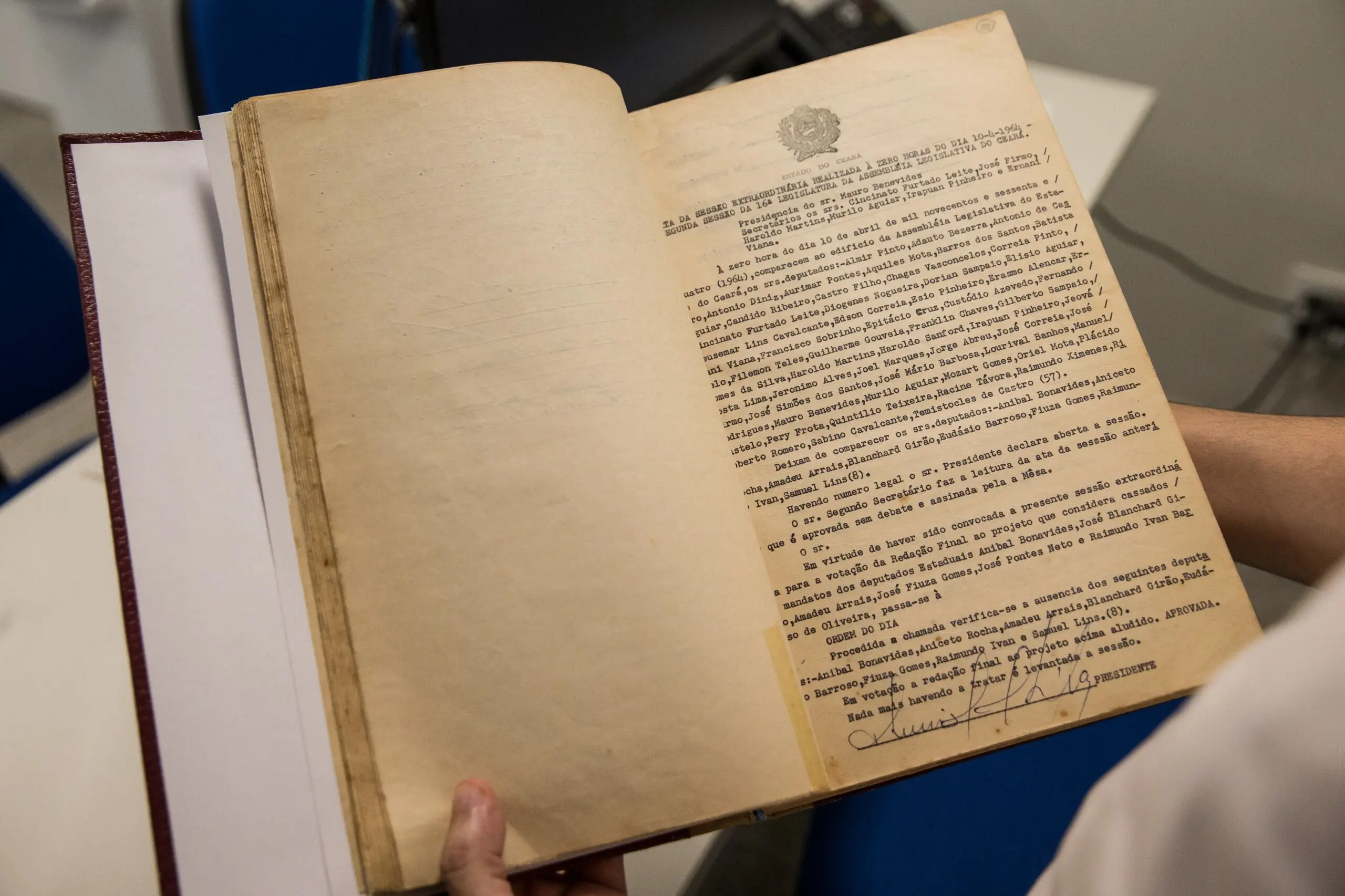 Imagem mostra o livre de atas da Assembleia, de 1964. A página em destaque é a do dia 10 de abril, em que deputados foram cassados. Um homem branco segura o livro para que o registro fotográfico seja feito. Na imagem, aparece parte de sua blusa branca e suas mãos, apenas.