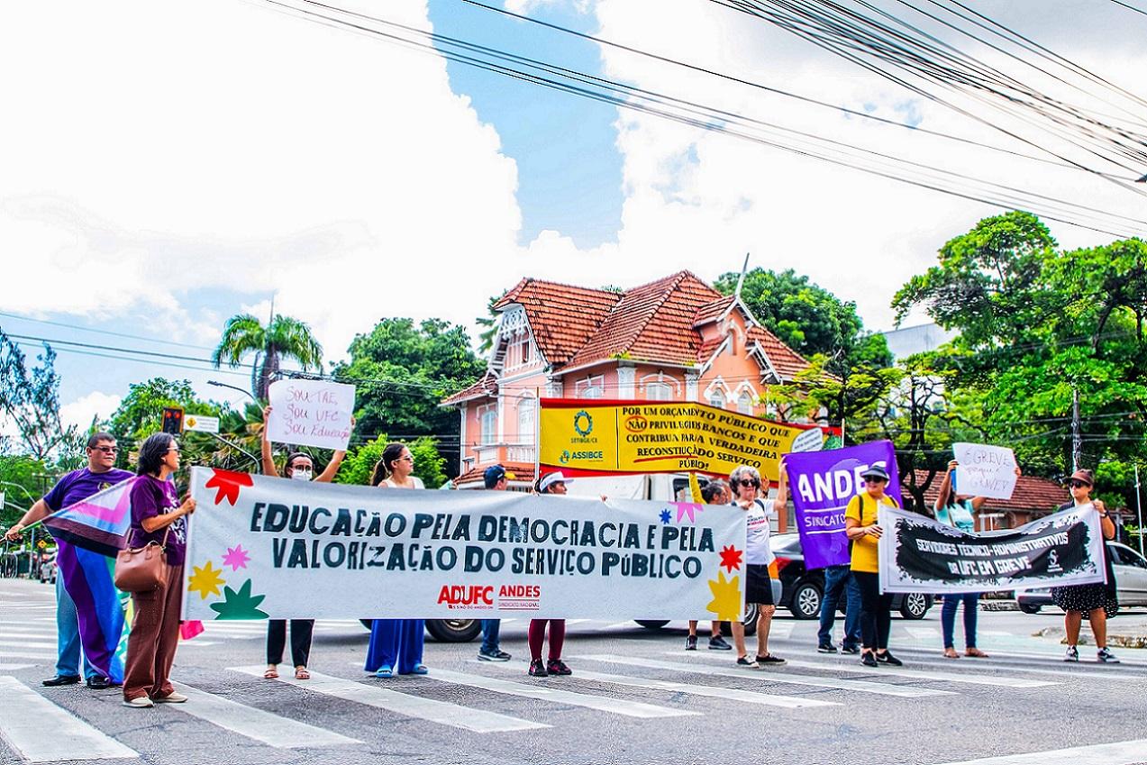 Servidores ocupam faixa de trânsito próxima à UFC com cartazes. Um deles diz: 'Educação pela democracia e pela valorização do serviço público'