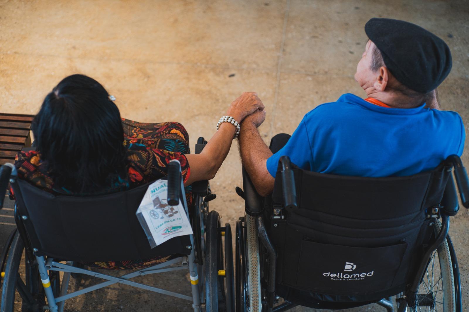 Sérgio e Rosália de mãos dadas, sentados cada um em sua cadeira de rodas