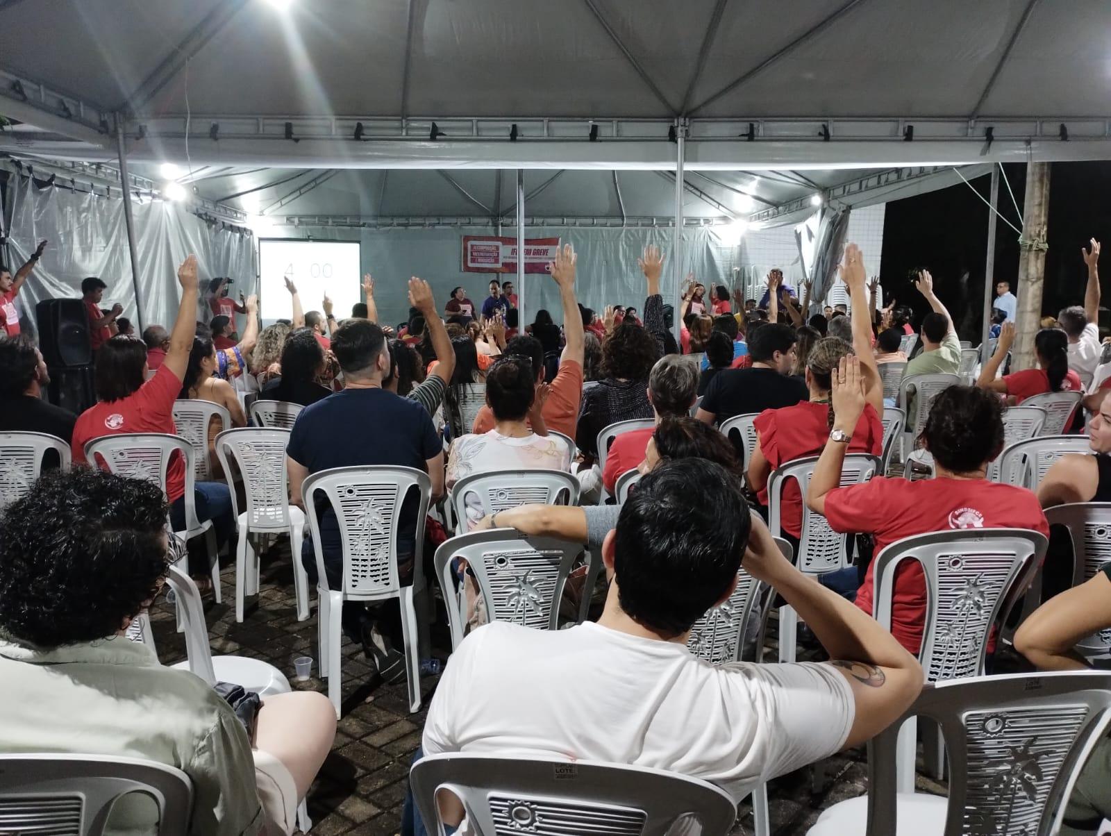 Pessoas de costas, sentadas em cadeiras. Parte delas está com braço levantado, votando em assembleia