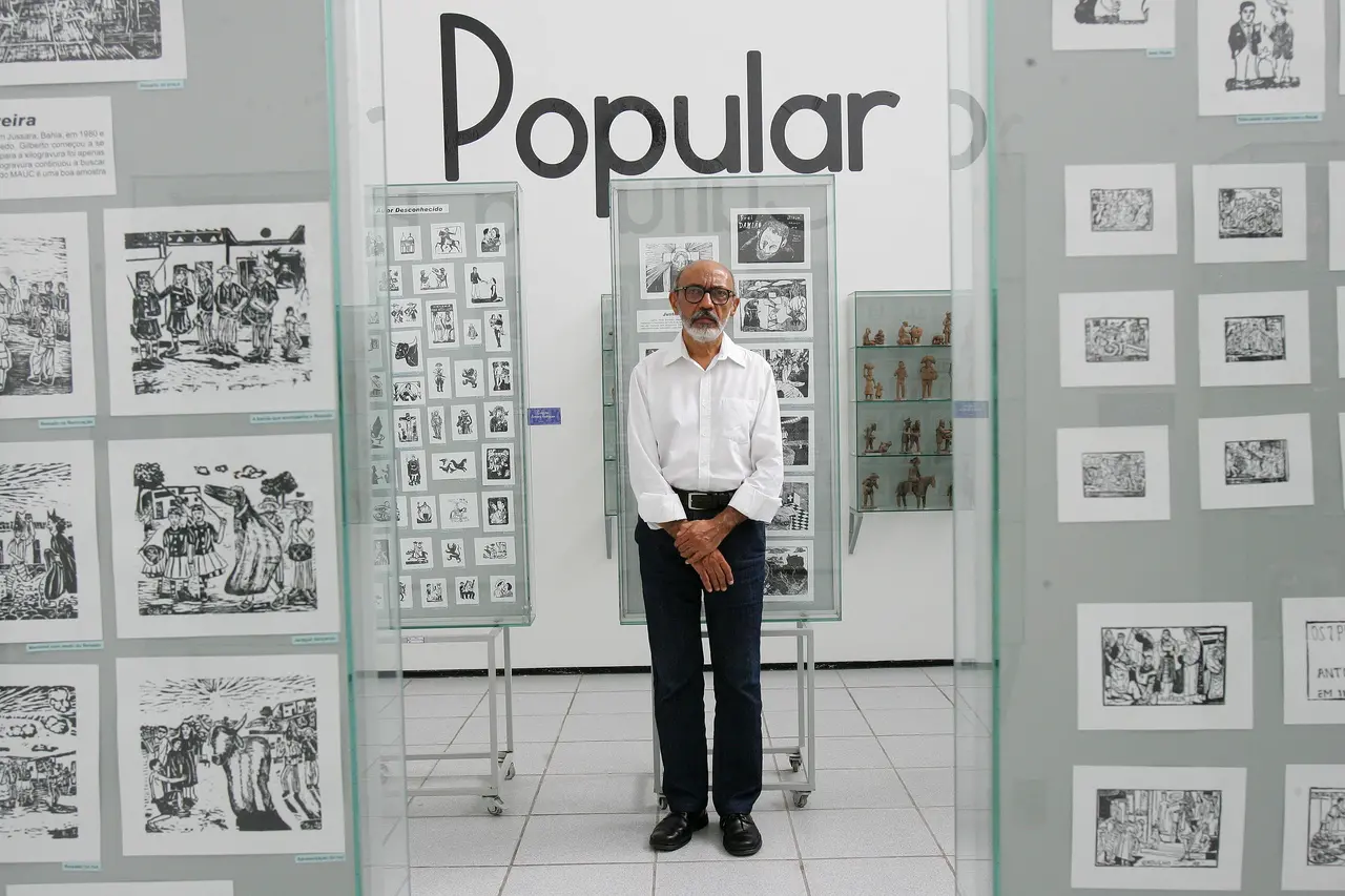 Gilmar de Carvalho  posa em frente a obras expostas em museu. Na parede atrás dele, a palavra 