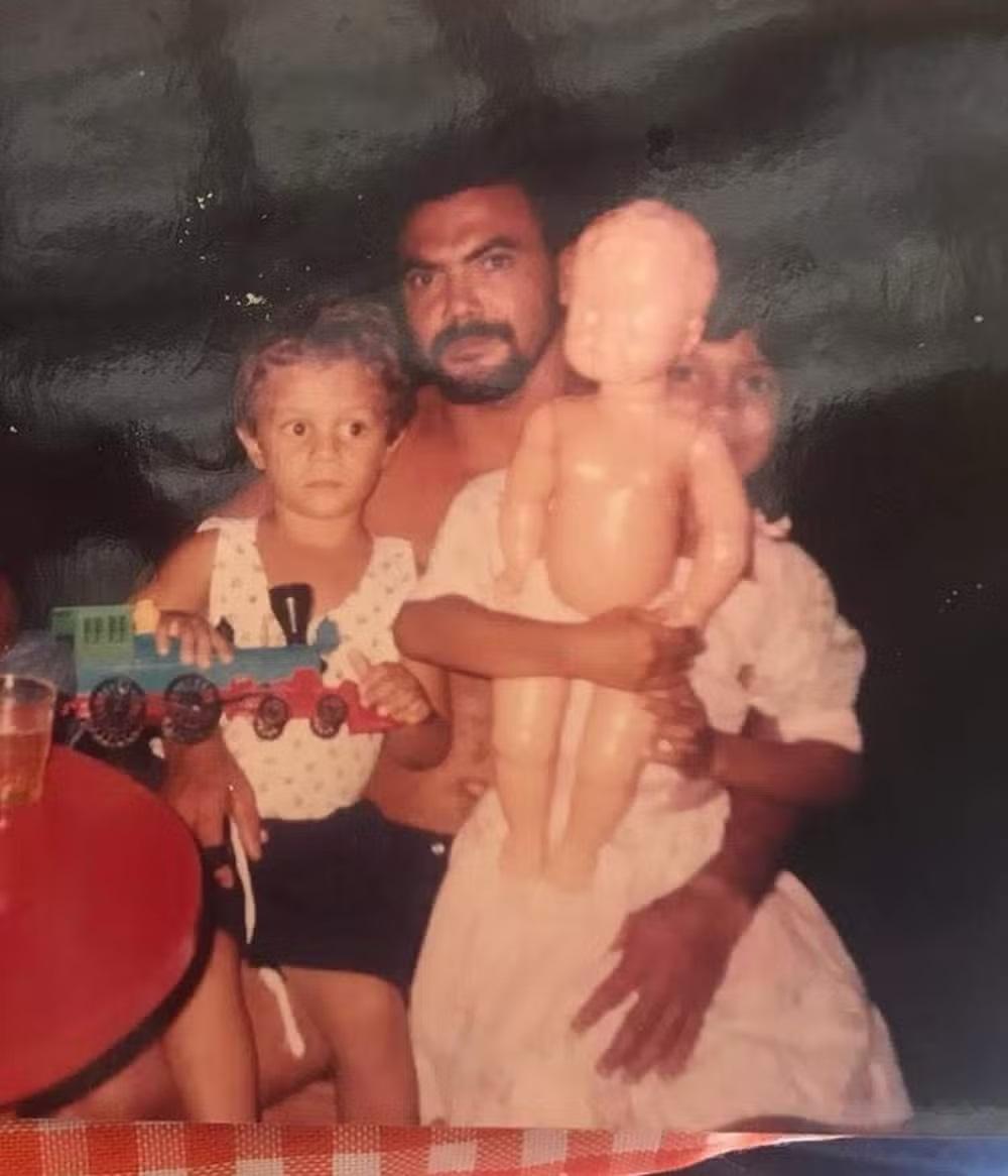 Gislayne, quando era pequena, aparece na foto segurando uma boneca, ao lado do pai e de outra criança