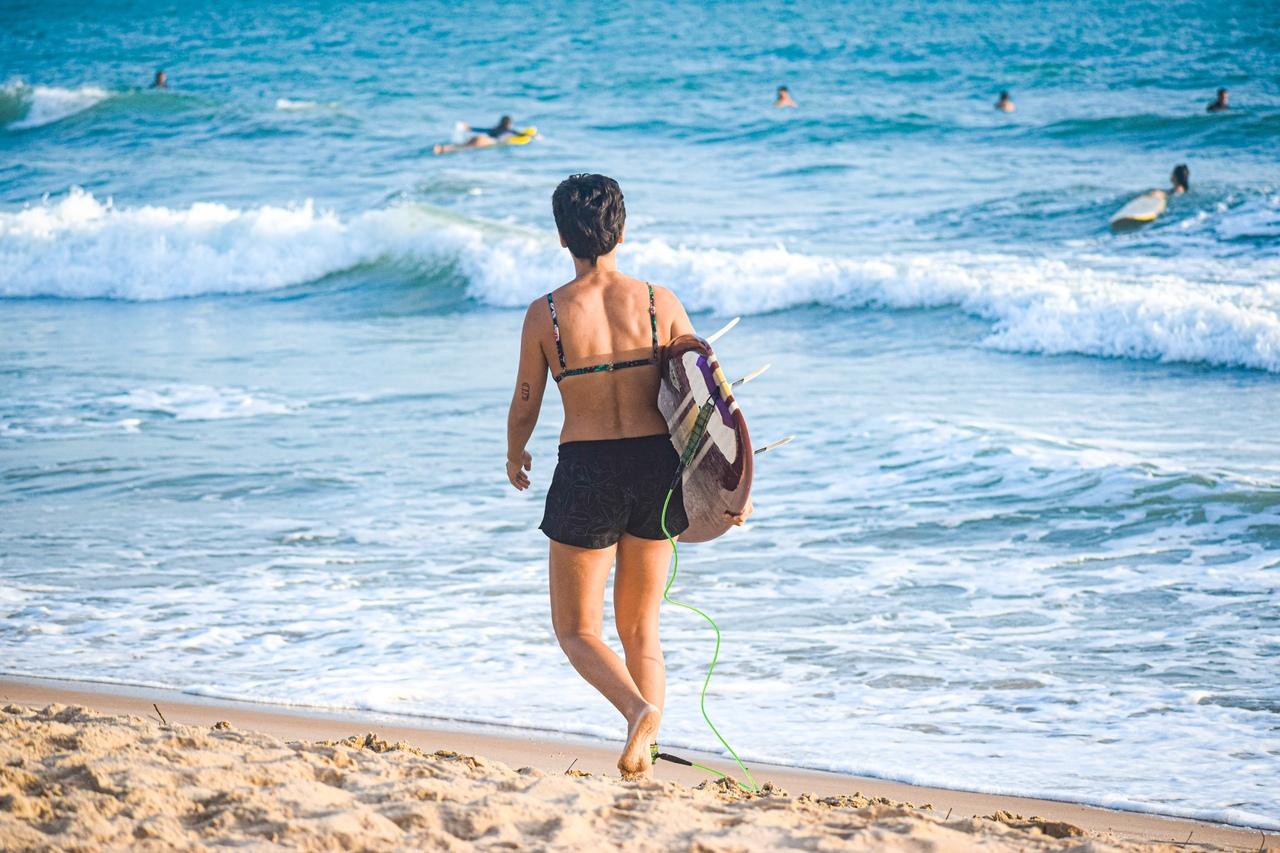 Mulher de costas, segurando uma prancha de surf, indo em direção ao mar