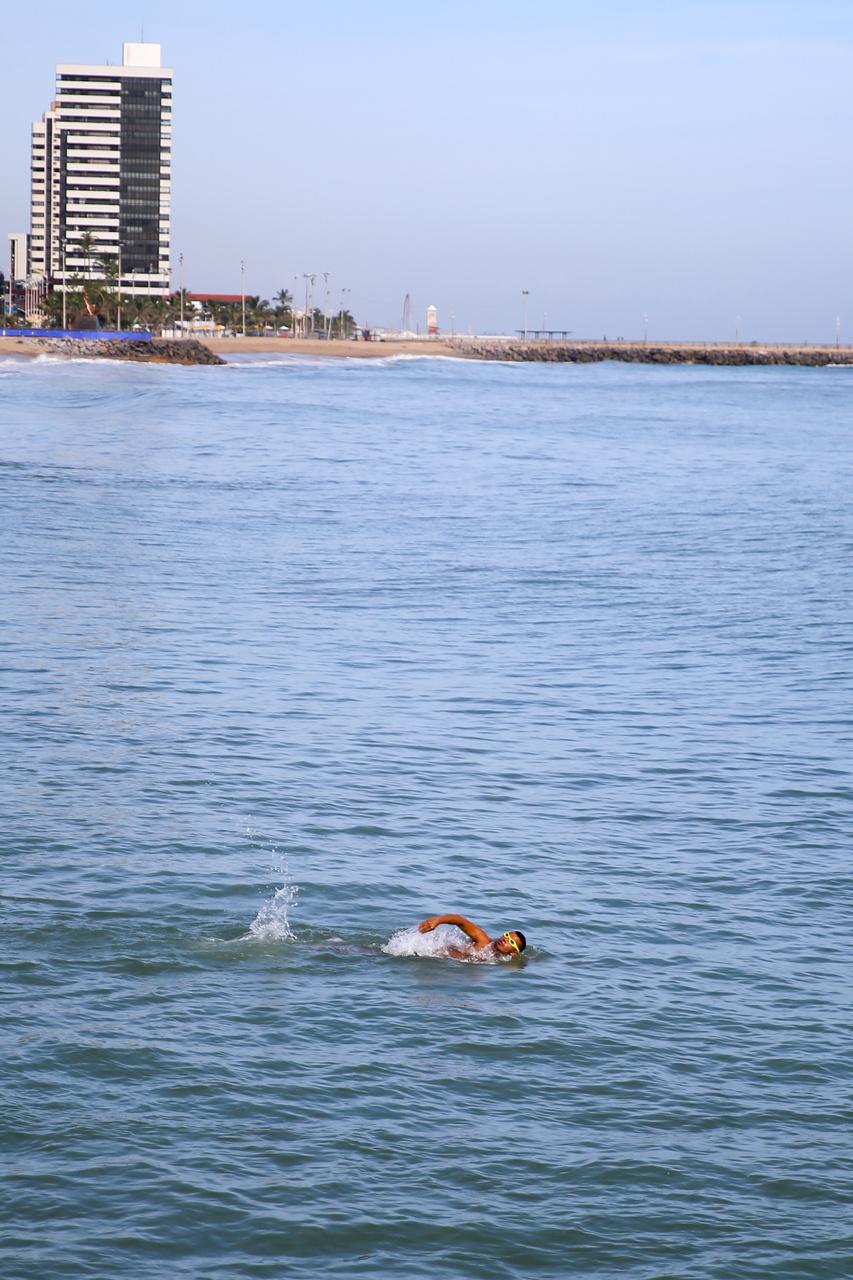 Pessoa praticando natação no mar. Em primeiro plano está um homem nadando e ao fundo aparece parte dos prédios da orla de Fortaleza