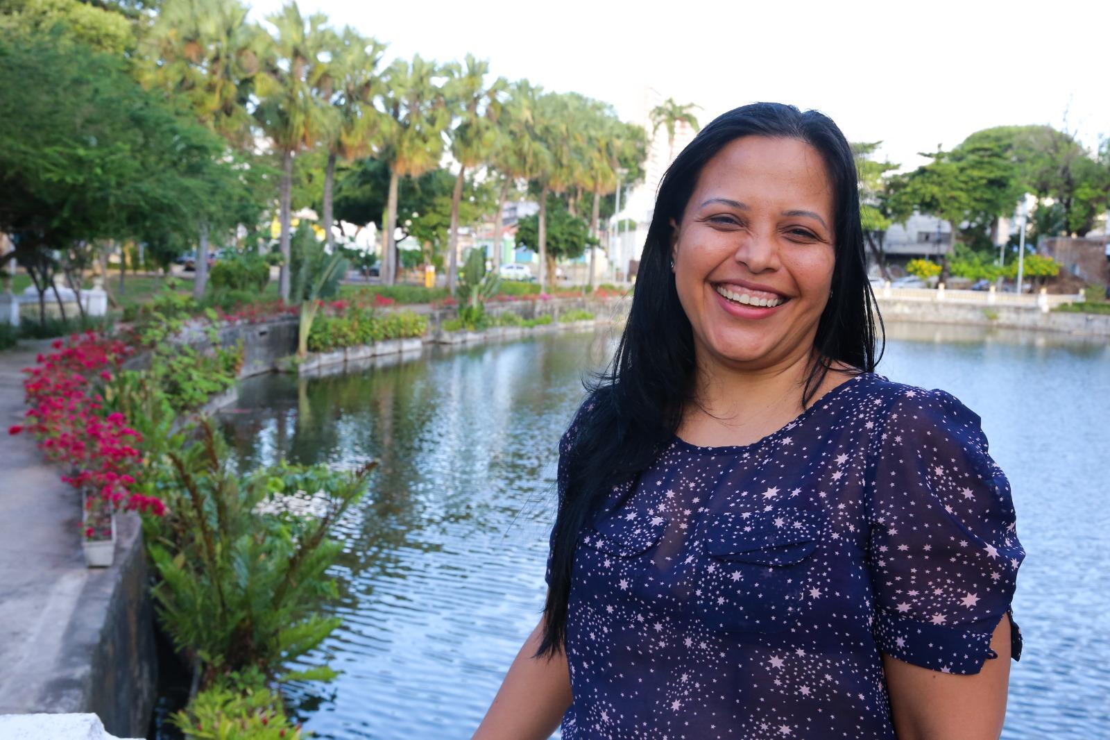 Eimily, mulher venezuelana de cabelos escuros e lisos, posa para foto sorrindo. Ao fundo, um lago e um parque de Fortaleza