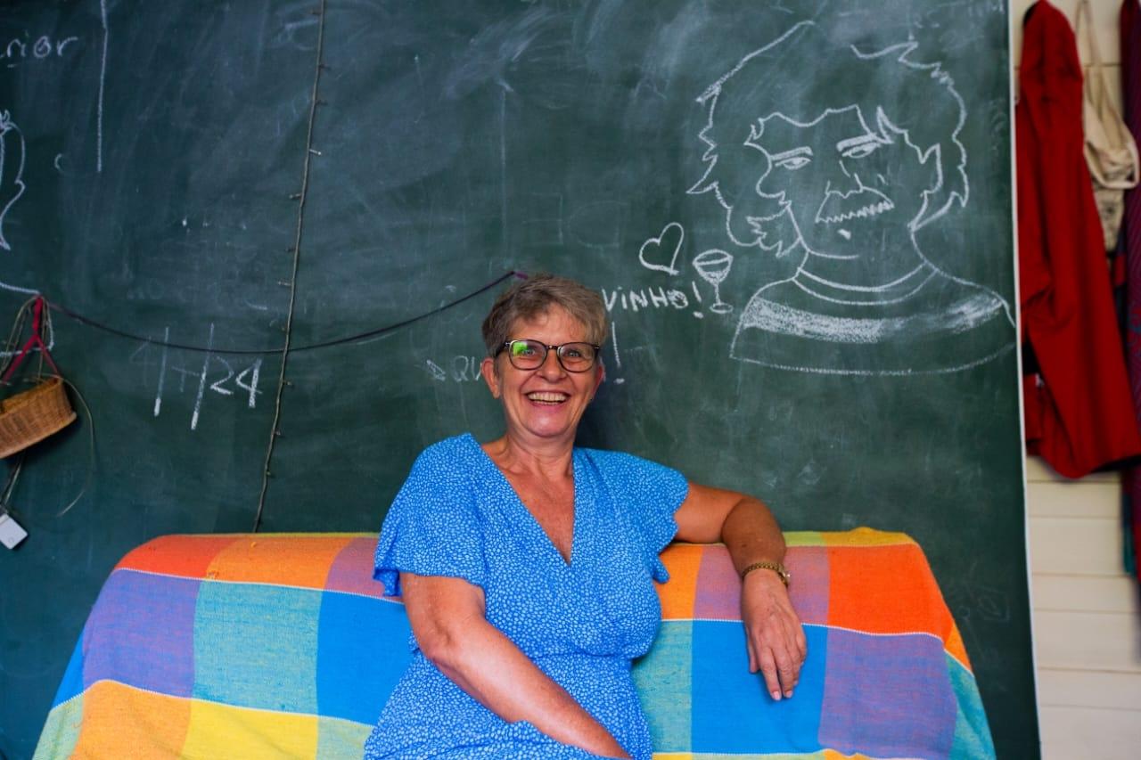 Rosina, branca e loira, de vestido azul. Ela está sentada, com o braço apoiado no sofá, sorrindo e olhando para a câmera