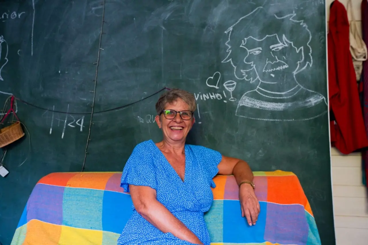 Rosina, branca e loira, de vestido azul. Ela está sentada, com o braço apoiado no sofá, sorrindo e olhando para a câmera