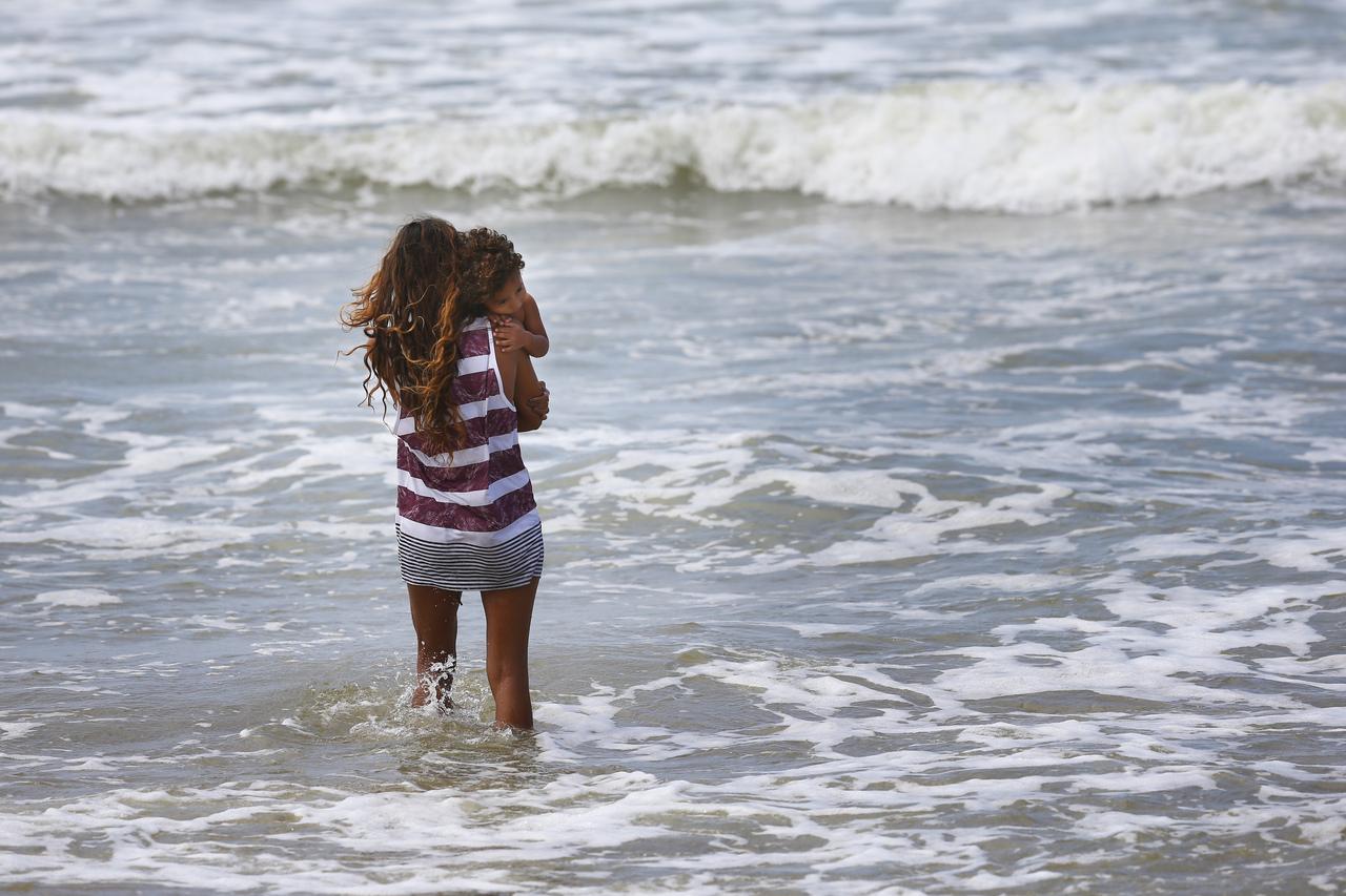 Mulher jovem segura criança no colo enquanto observa o mar, com as águas até metade da perna