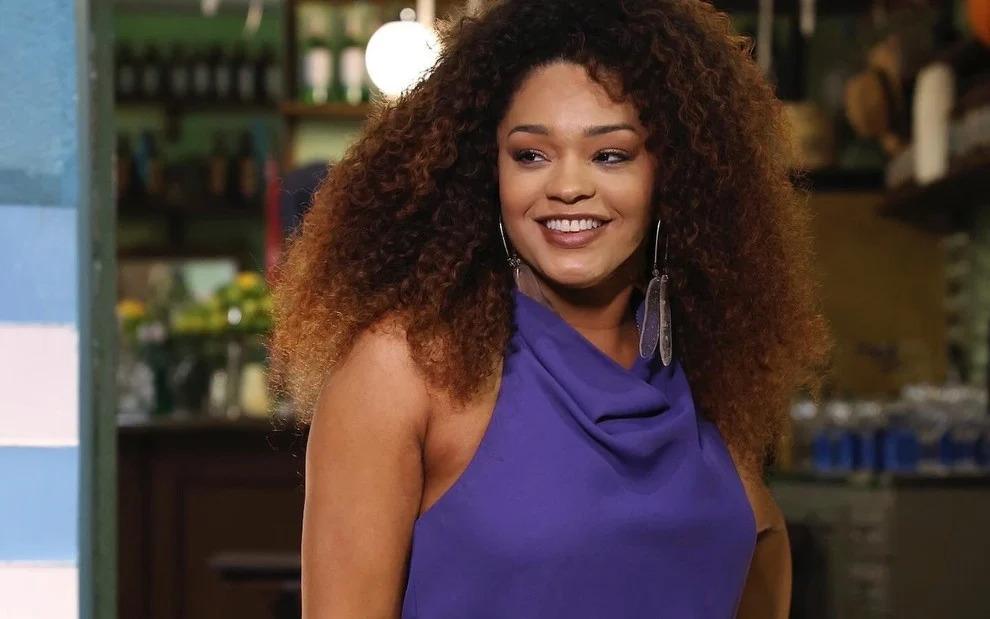 Juliana Alves Imagem de uma jovem mulher com cabelo cacheado e volumoso, sorrindo em um ambiente descontraído. Ela veste uma blusa roxa, exibindo um estilo alegre e confiante.