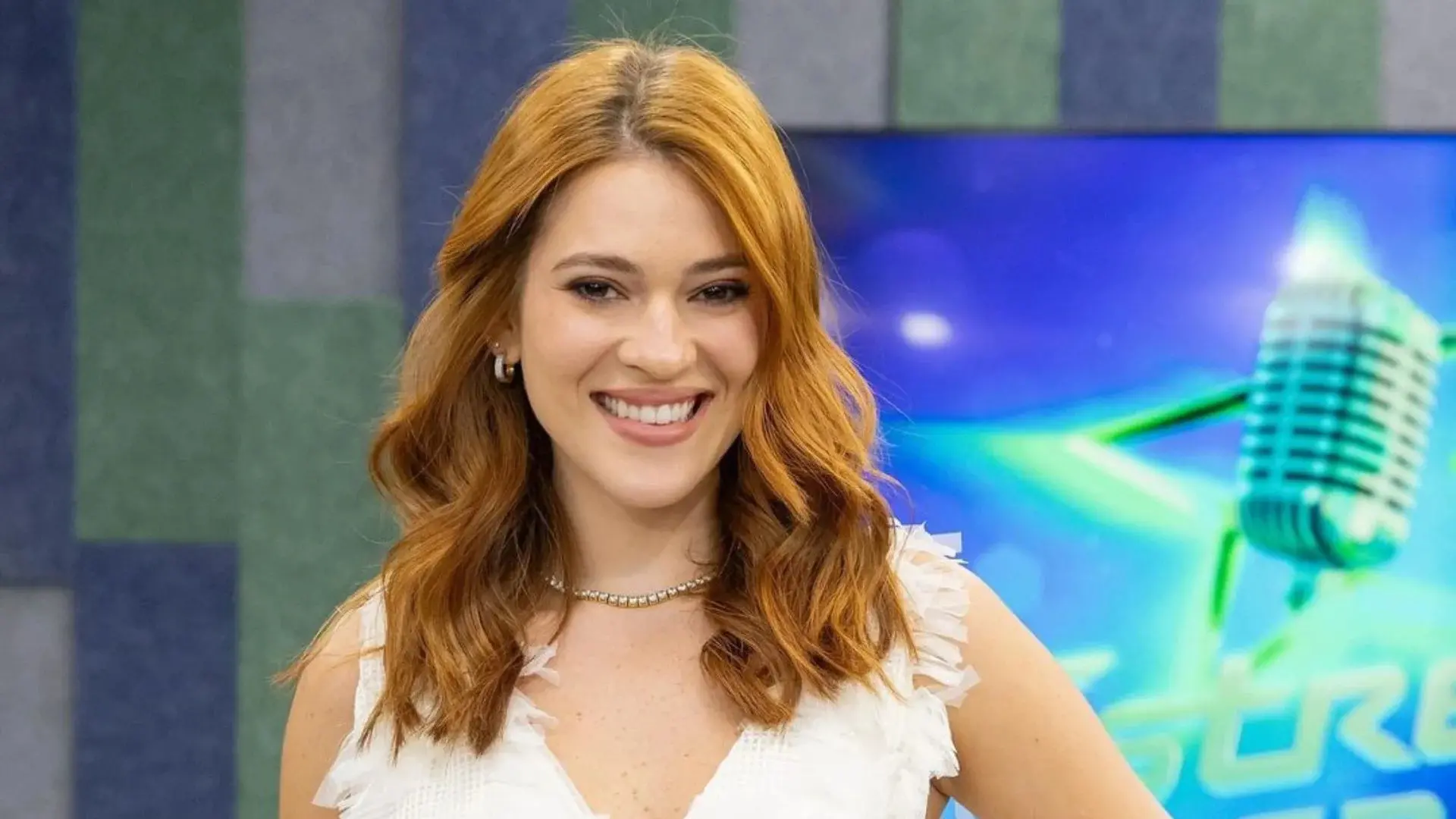 Imagem de uma mulher sorrindo com cabelos ondulados e ruivos, em um cenário de estúdio com elementos de música. Ela usa uma blusa branca e acessórios elegantes.