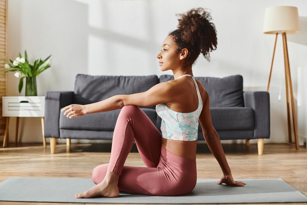 Mulher negra de cabelo cacheado amarrado em um coque praticando yoga. Ela usa top branco e calça rosé