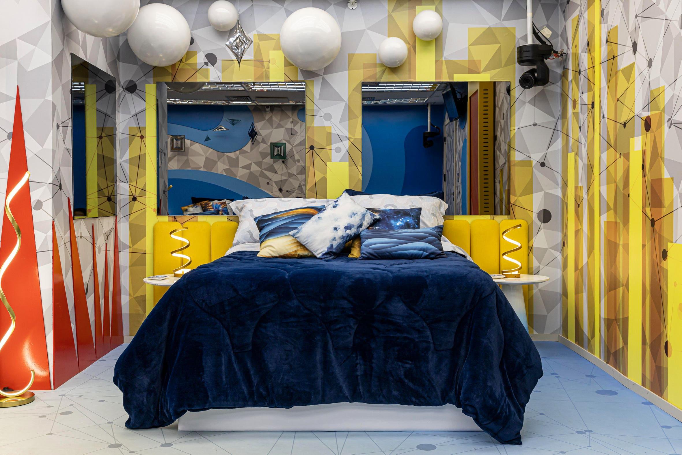 Quarto decorado com cama azul e almofadas coloridas, paredes geométricas em tons de amarelo e azul, criando um ambiente contemporâneo e vibrante.