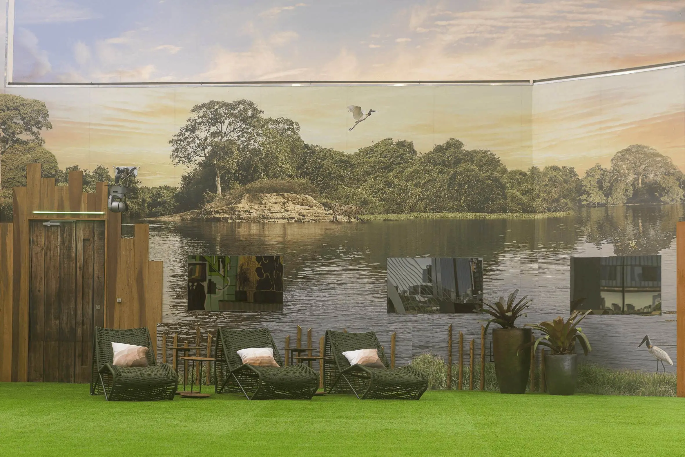 Imagem de um espaço interno com decoração que remete à natureza, incluindo um mural de paisagem com árvore e lago, cadeiras confortáveis e plantas.