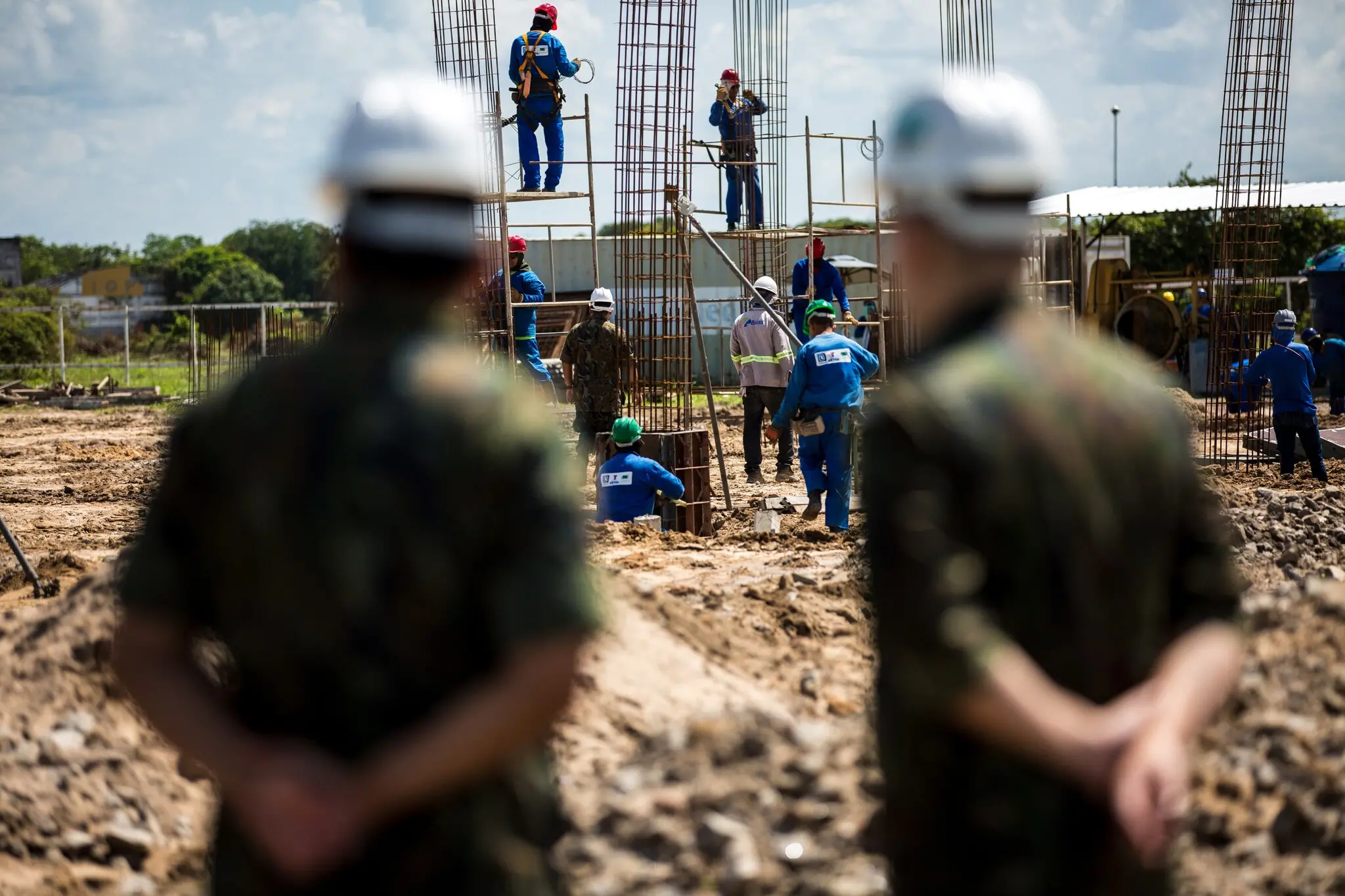 Vista do canteiro de obras ao fundo, em foco, com dois militares em primeiro plano, desfocados, observando o trabalho