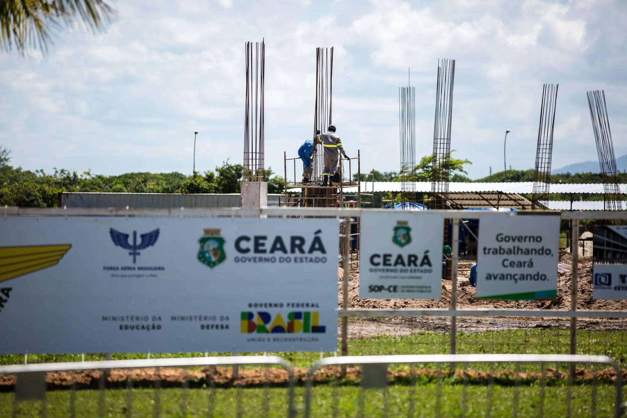 Construção em andamento com trabalhadores sobre um andaime, estruturas metálicas erguidas e placas indicando apoio do governo do Ceará e do governo federal.