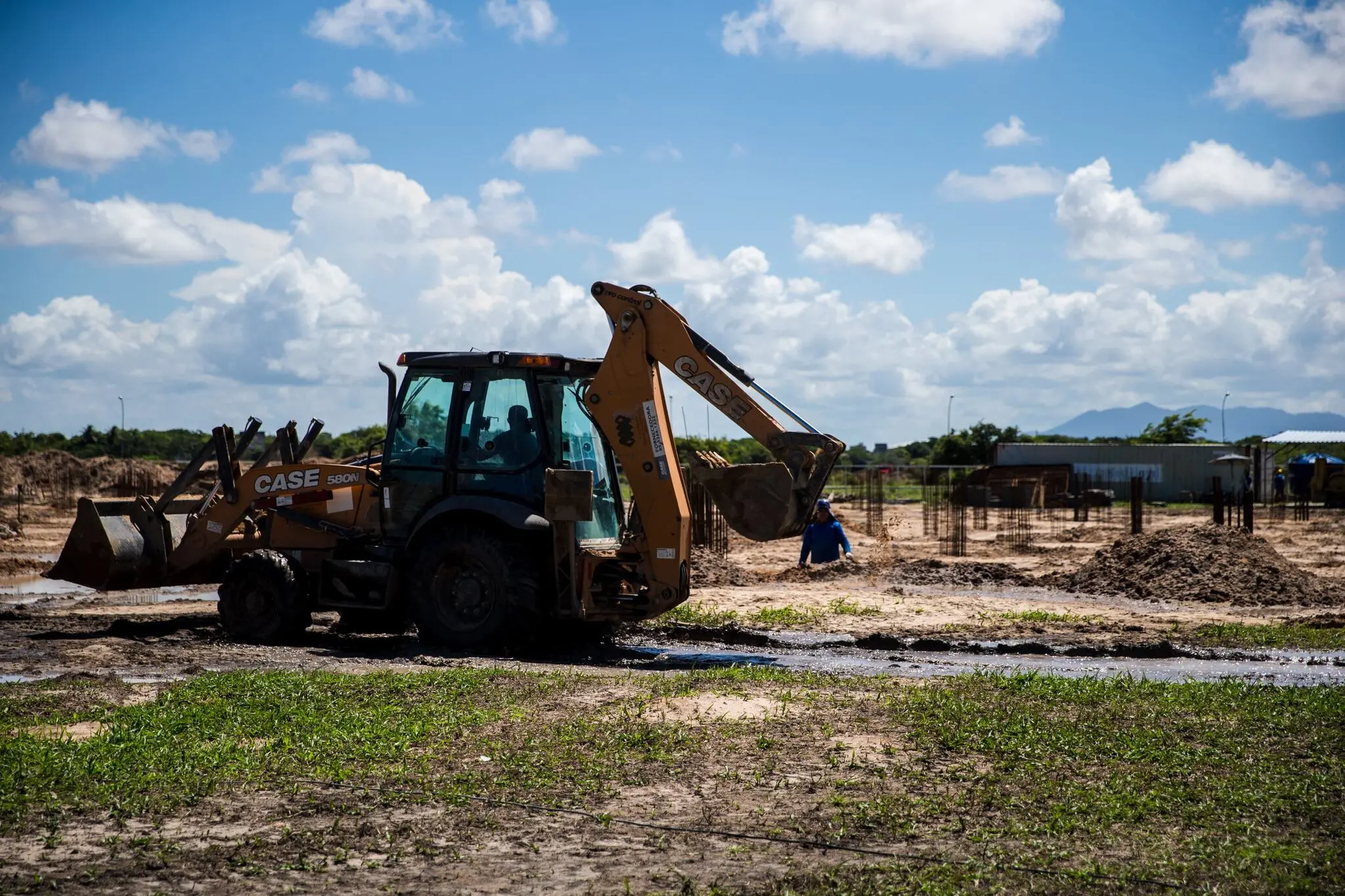 Retroescavadeira operando no terreno de construção, com trabalhador ao fundo lidando com a fundação do edifício.