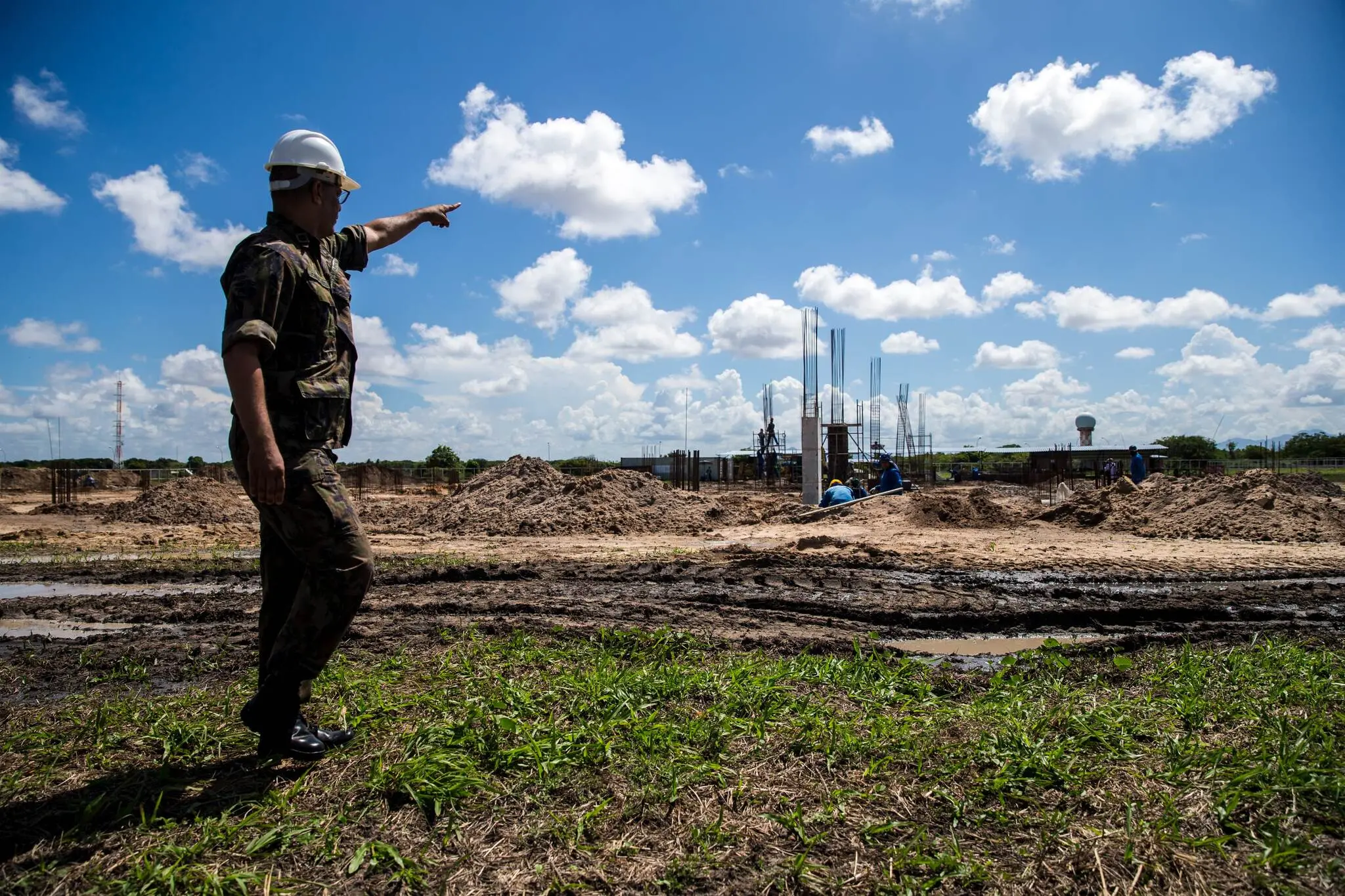 Um homem usando capacete branco e uniforme camuflado aponta para o horizonte em um canteiro de obras, com estruturas de ferro ao fundo. O terreno é de terra.