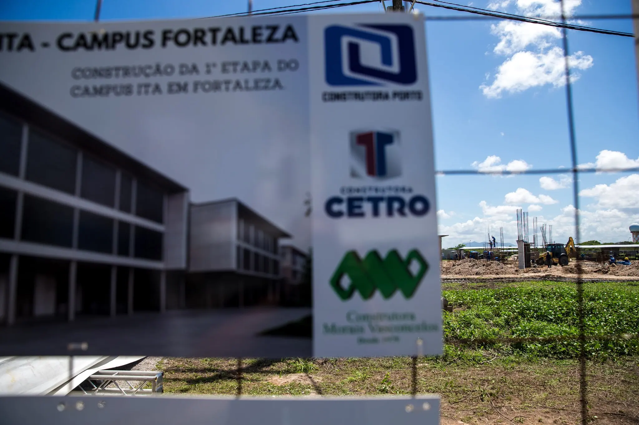 Construção em andamento com trabalhadores sobre um andaime, ao fundo, com estruturas metálicas erguidas e placas indicando a construção da 1ª etapa do campus ITA em Fortaleza