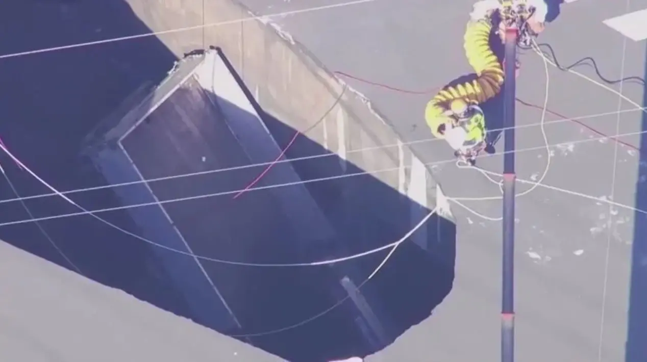 Imagem aérea mostrando um buraco grande em uma estrada com um trabalhador utilizando equipamento de segurança próximo a fios elétricos. Perigo de deslizamento de terra.