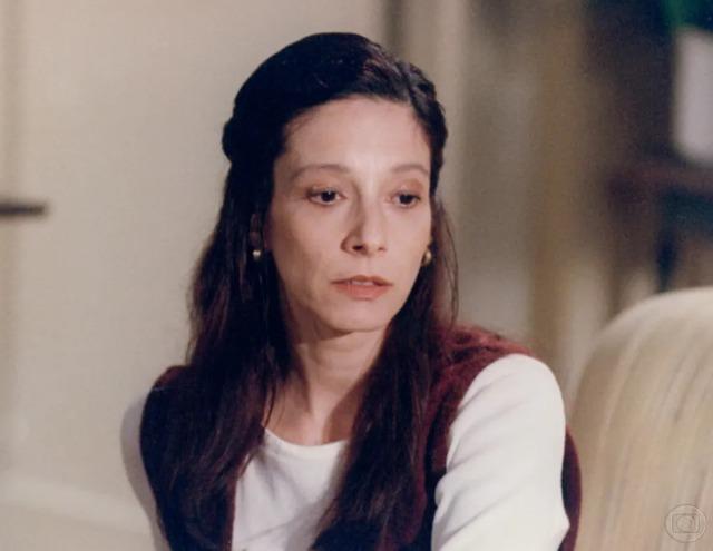 Mulher com cabelo longo e castanho, usando uma blusa branca e colete marrom, em ambiente interno, demonstrando expressão pensativa.