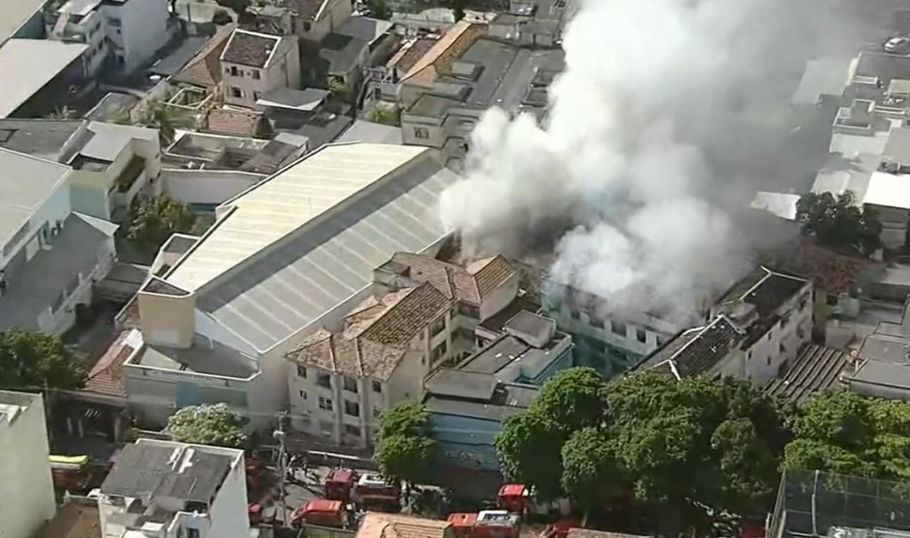 Imagem de um incêndio em um edifício, mostrando a fumaça saindo do telhado em uma área urbana, com bombeiros presentes na cena.