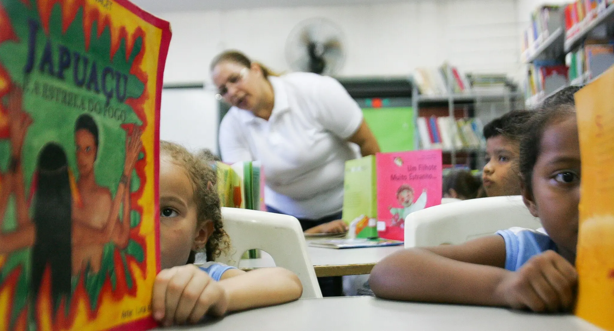Crianças em uma biblioteca, segurando livros coloridos. Em primeiro plano, duas meninas aparecem com os rostos parcialmente cobertos pelos livros que estão lendo.