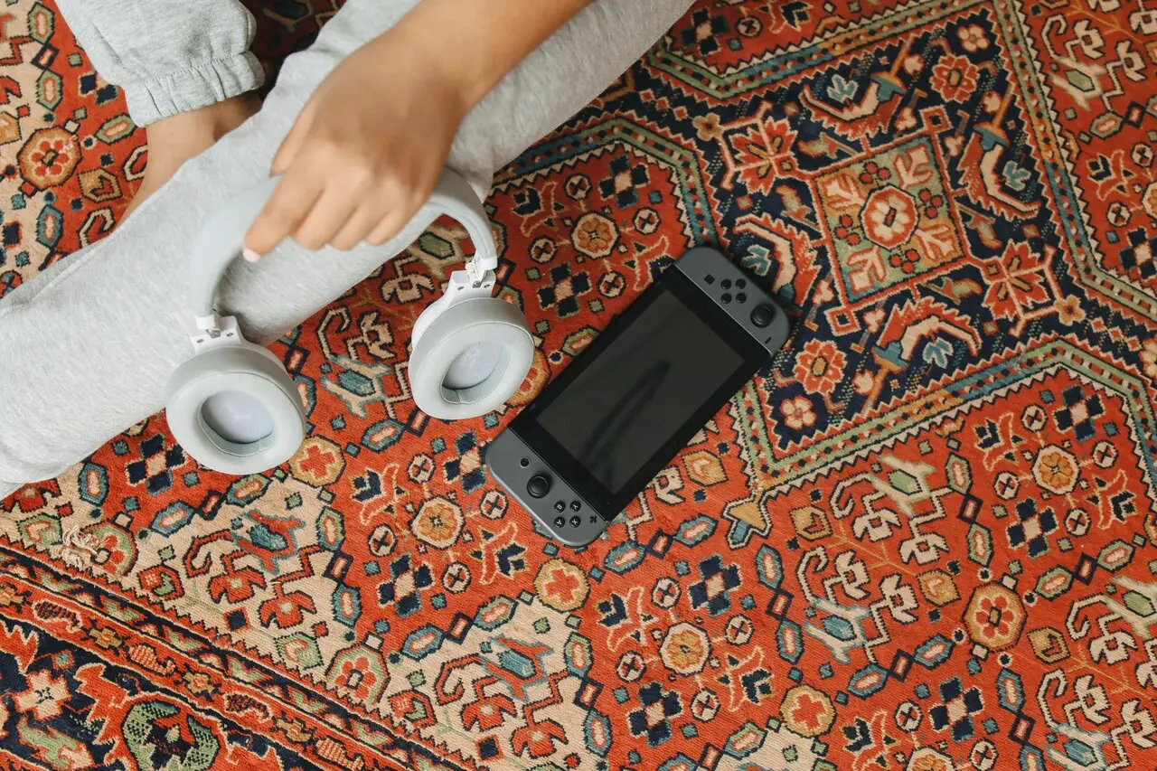 Mão segurando fone de ouvido branco ao lado de um celular em cima de uma tapeçaria