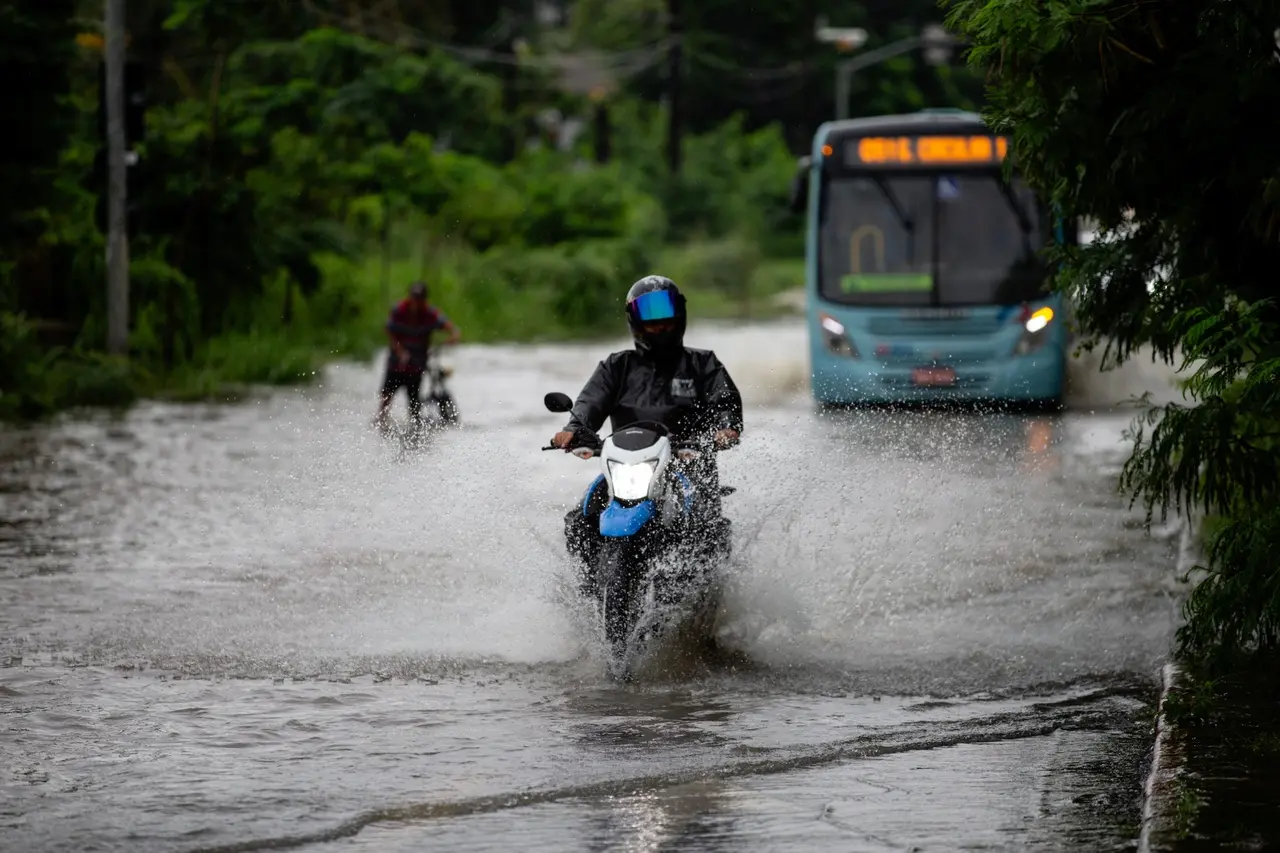 Foto de rua alagada durante chuva em Fortaleza. Na foto, aparecem um motoqueiro e um homem em uma bicicleta e um ônibus ao fundo