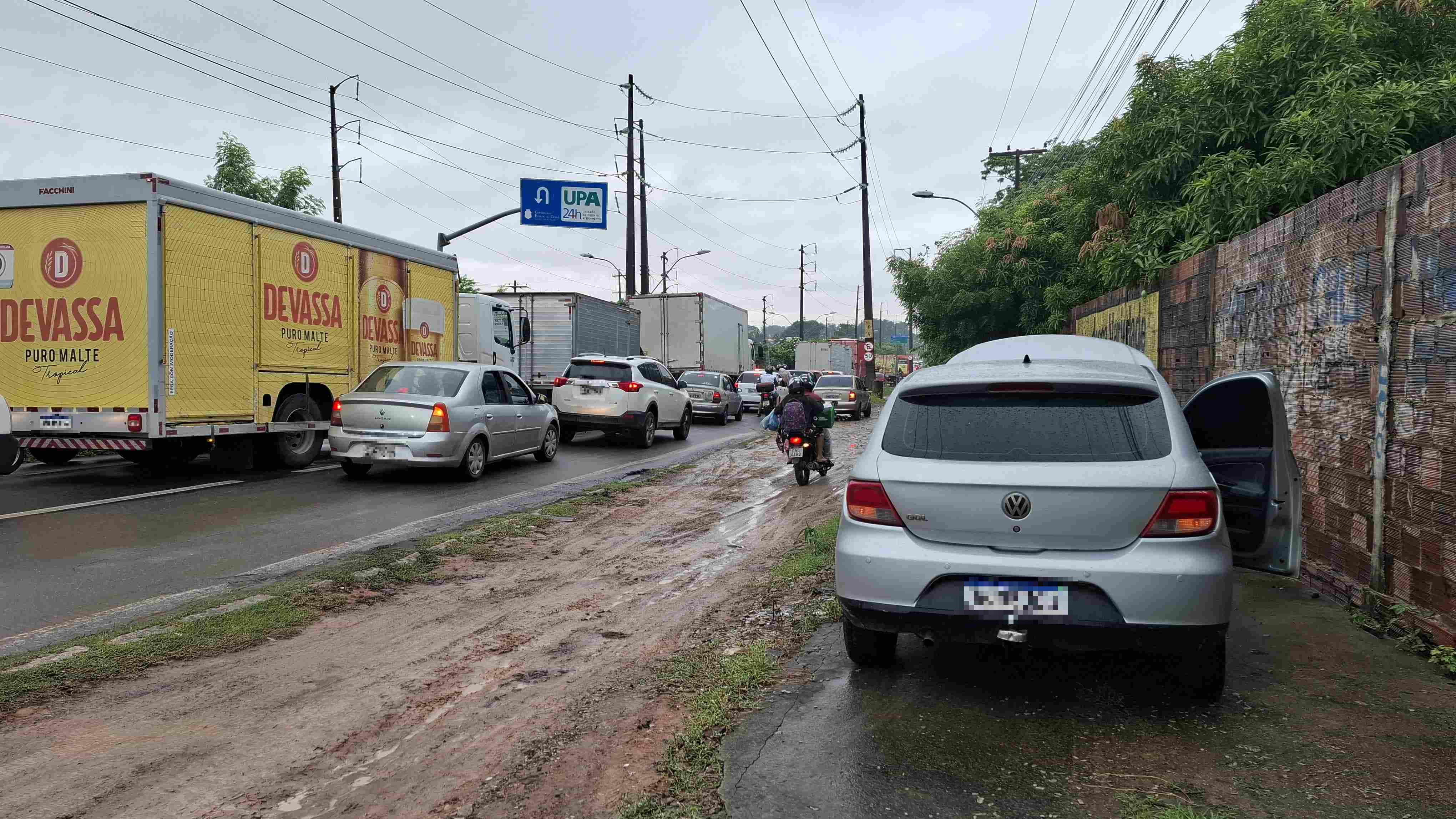Imagem mostrando um engarrafamento em uma via com diversos veículos, incluindo carros e caminhões, e um anúncio de UPA ao fundo.