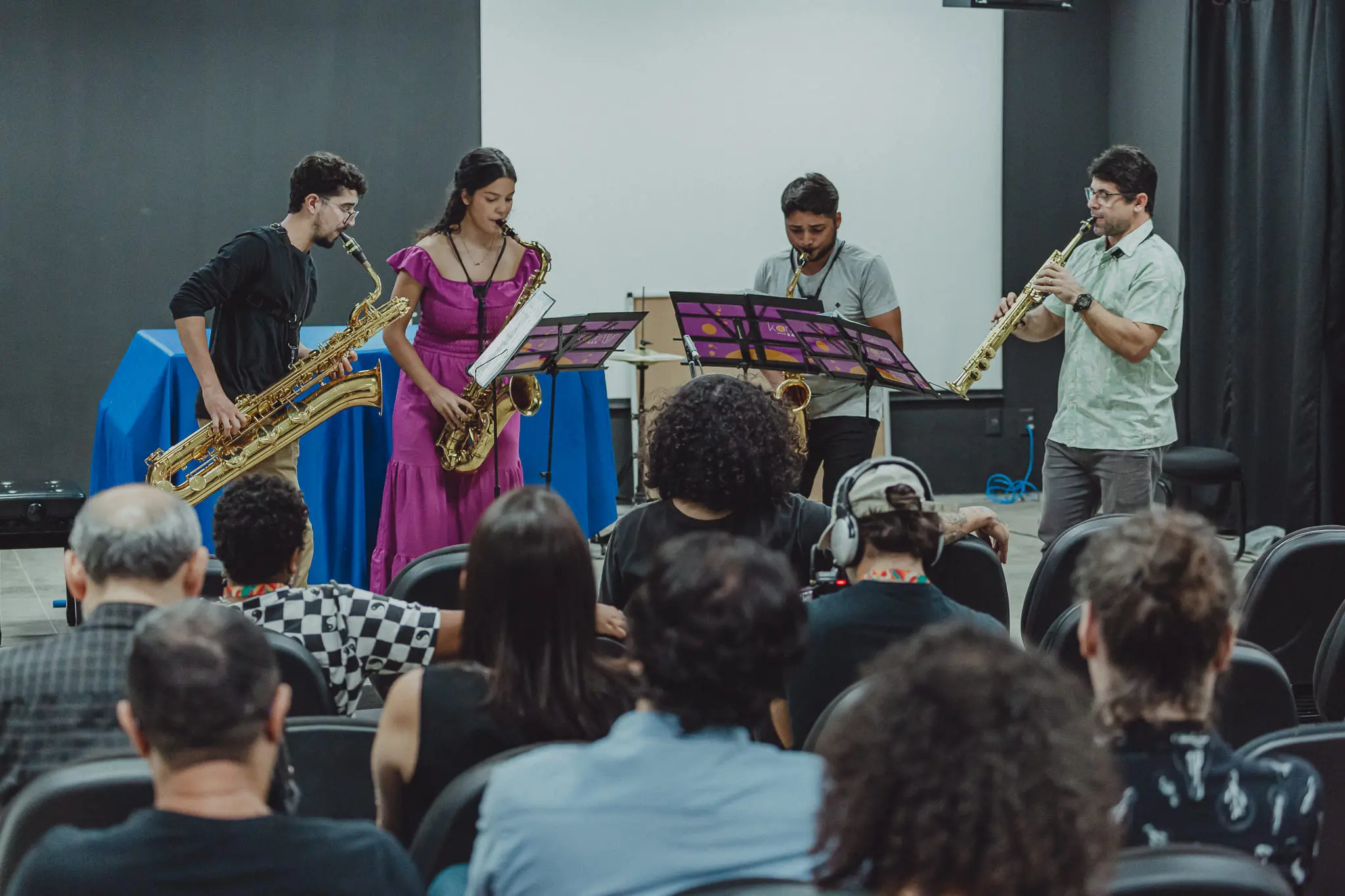 Apresentação musical com quatro músicos tocando saxofones em um auditório. O público acompanha atentamente a performance ao vivo.