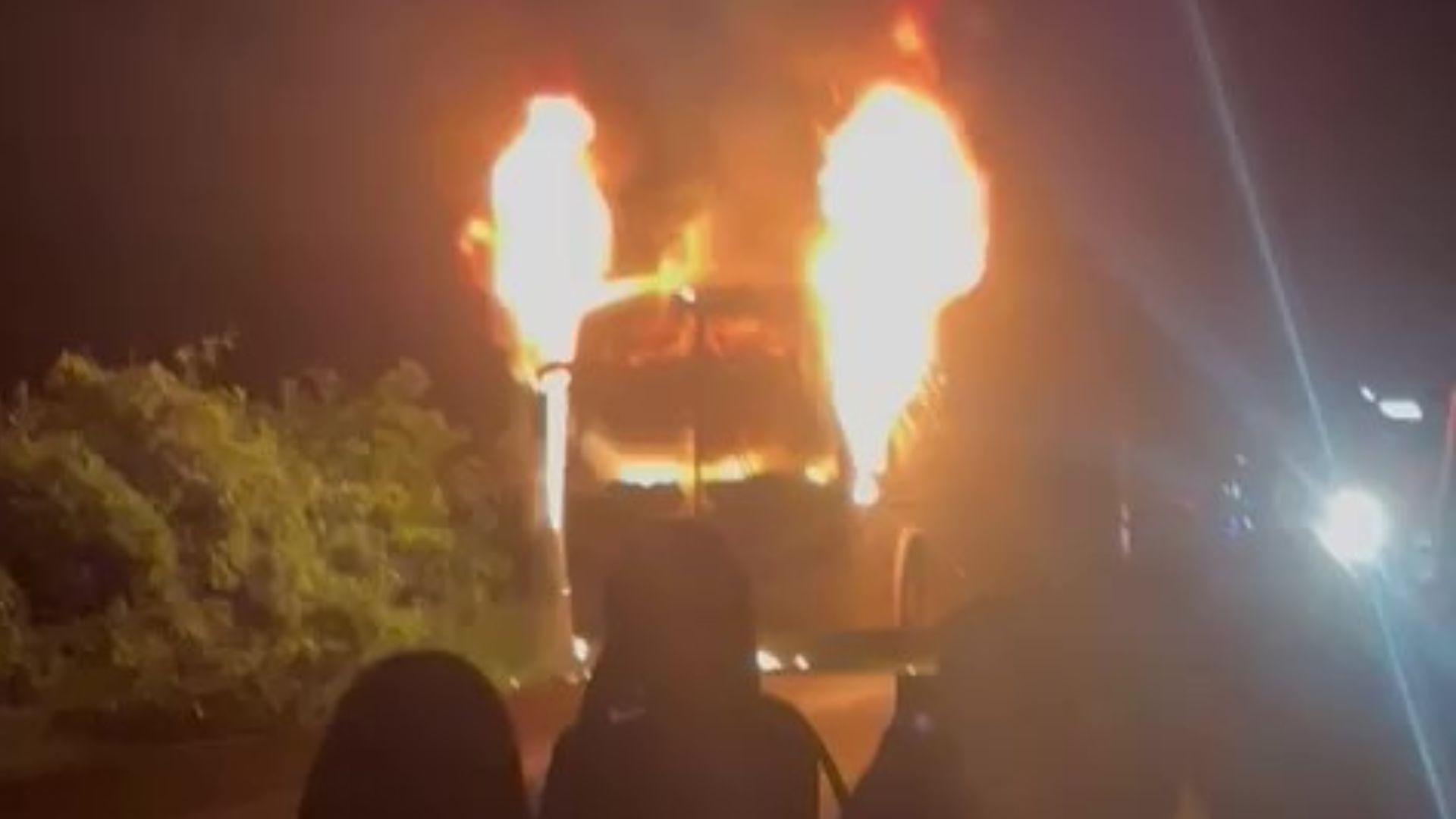 Ônibus em chamas à noite, com fogo saindo da parte frontal e espectadores ao fundo, de forma impressionante e impactante.