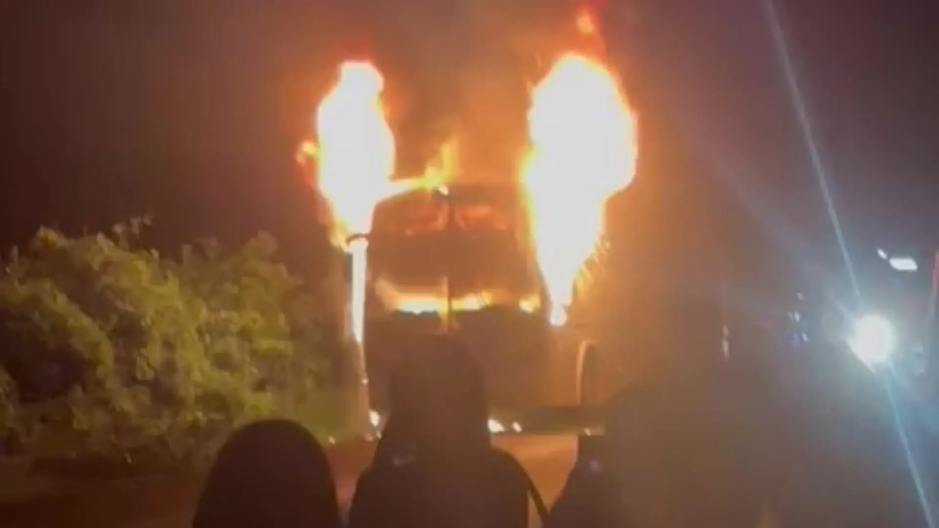 Ônibus em chamas à noite, com fogo saindo da parte frontal e espectadores ao fundo, de forma impressionante e impactante.