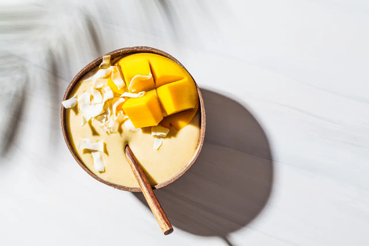 Uma tigela de smoothie cremoso com manga picada com banana batida e lascas de coco, perfeita para um lanche saudável e refrescante.