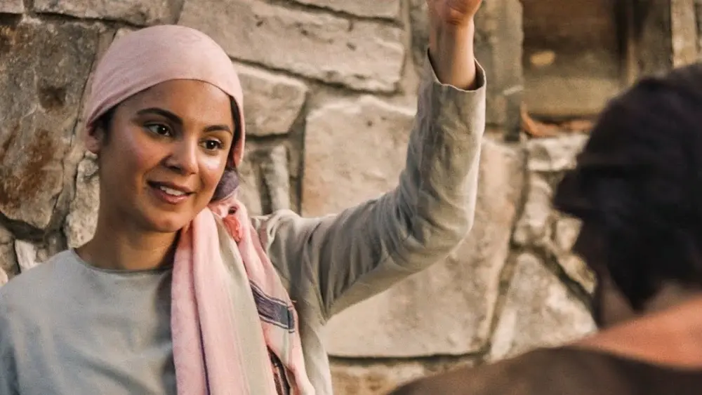 Mulher com lenço na cabeça acenando e sorrindo, em ambiente rústico, ideal para temas de cultura e tradições.