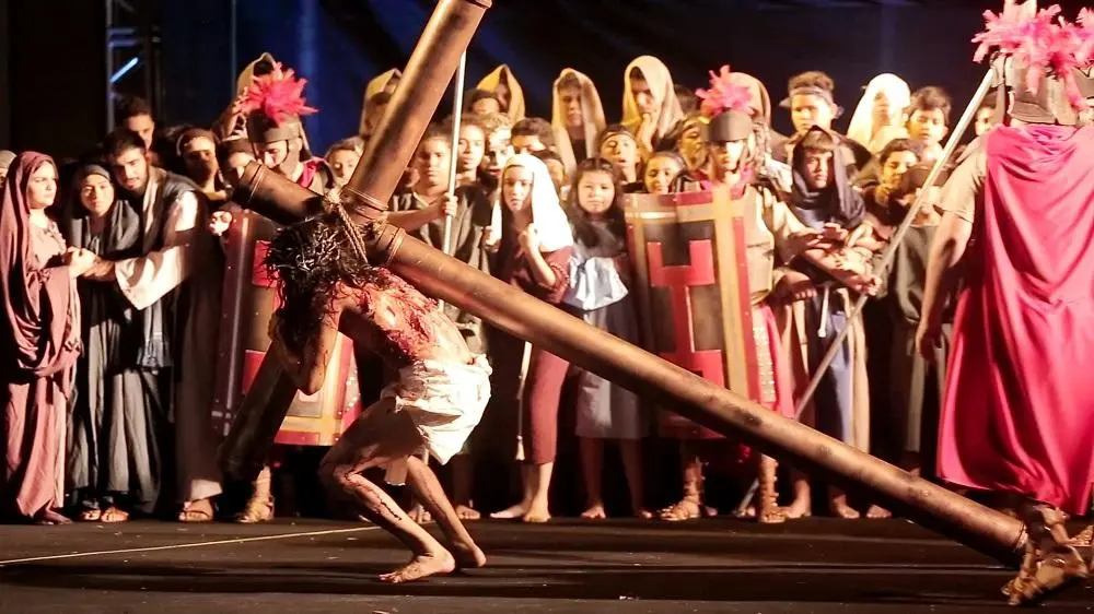 Imagem de cena da Paixão de Cristo, com Jesus carregando a cruz de madeira