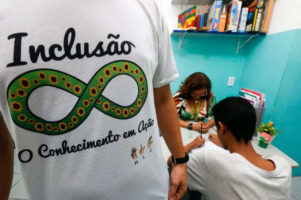 imagem com um busto de uma pessoa usando uma blusa com mensagem favorável à inclusão. Ao fundo já uma mulher e um estudante em volta de uma mesa de estudos.