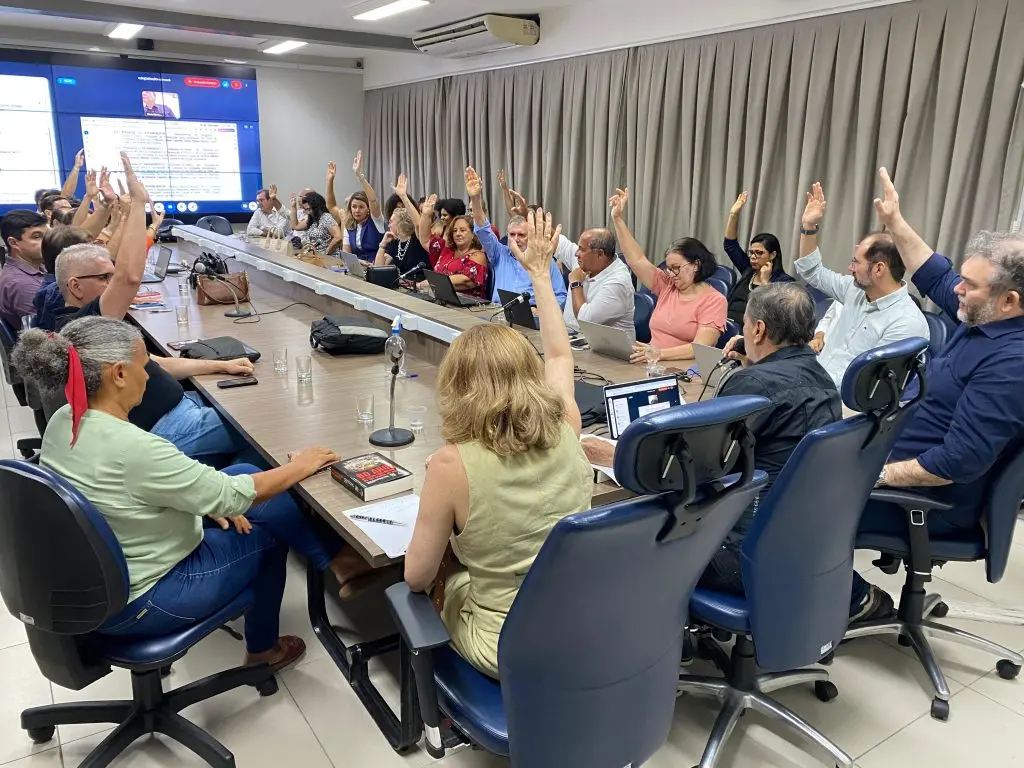 Imagem de integrantes do  Conselho Universitário da UFRN, com braços erguidos em votação em volta de uma mesa