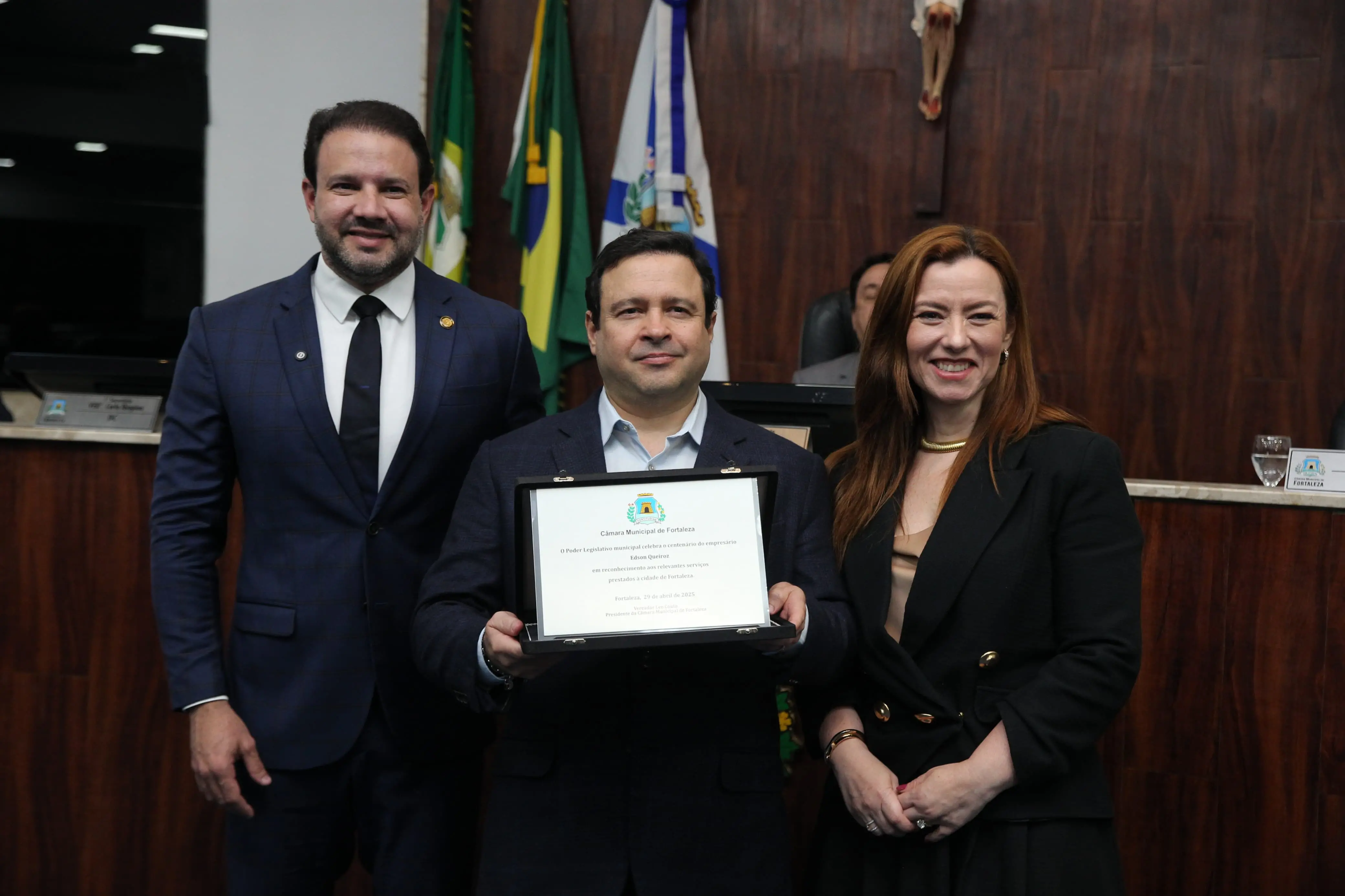 Foto do presidente da Câmara Municipal de Fortaleza Léo Couto, ao lado de Igor Queiroz Barroso e sua esposa, Aline Félix Barroso, no plenário da Câmara Municipal de Fortaleza. Na foto, Igor Queiroz Barroso segura uma placa em homenagem ao centenário de Edson Queiroz