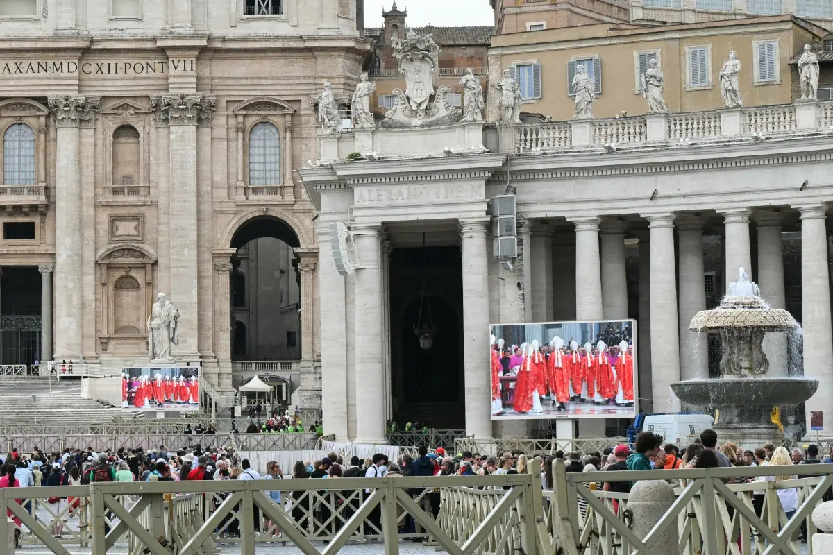 Vista da Praça de São Pedro, com multidão e telões exibindo atividades religiosas, retratando a importância cultural e espiritual do local.