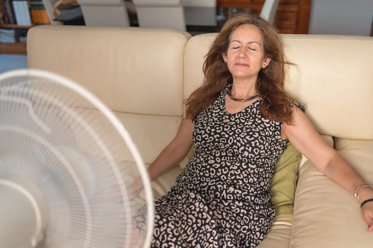Mulher mais velha com expressão cansada, sentada em um sofá, em frente ao ventilador, retratando os desafios emocionais e físicos enfrentados por muitas mulheres durante o climatério.