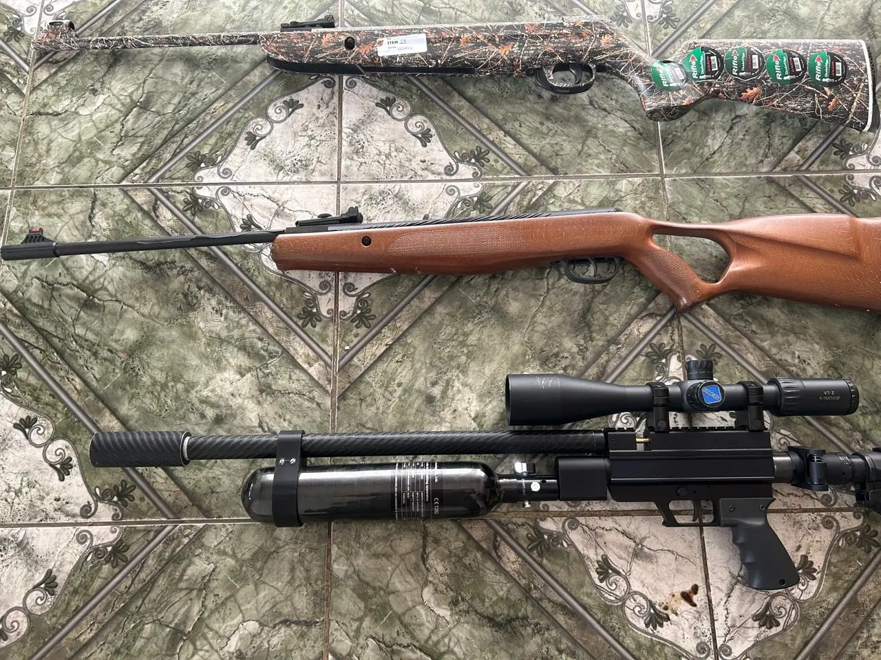 Imagem mostra duas armas de fogo, uma espingarda de madeira e um rifle com mira telescópica, em um ambiente com padrão decorativo de mármore.