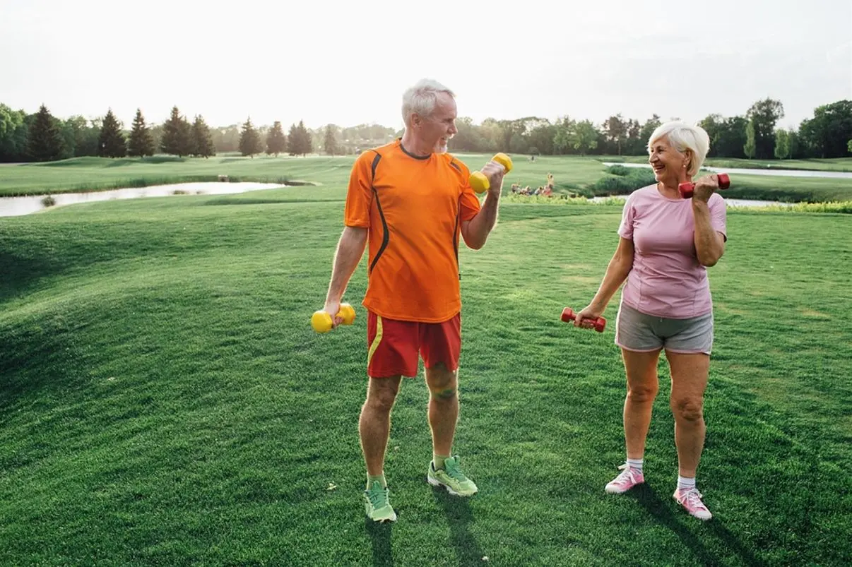 foto de dois idosos praticando atividade física sob a grama em parque cheio de árvores
