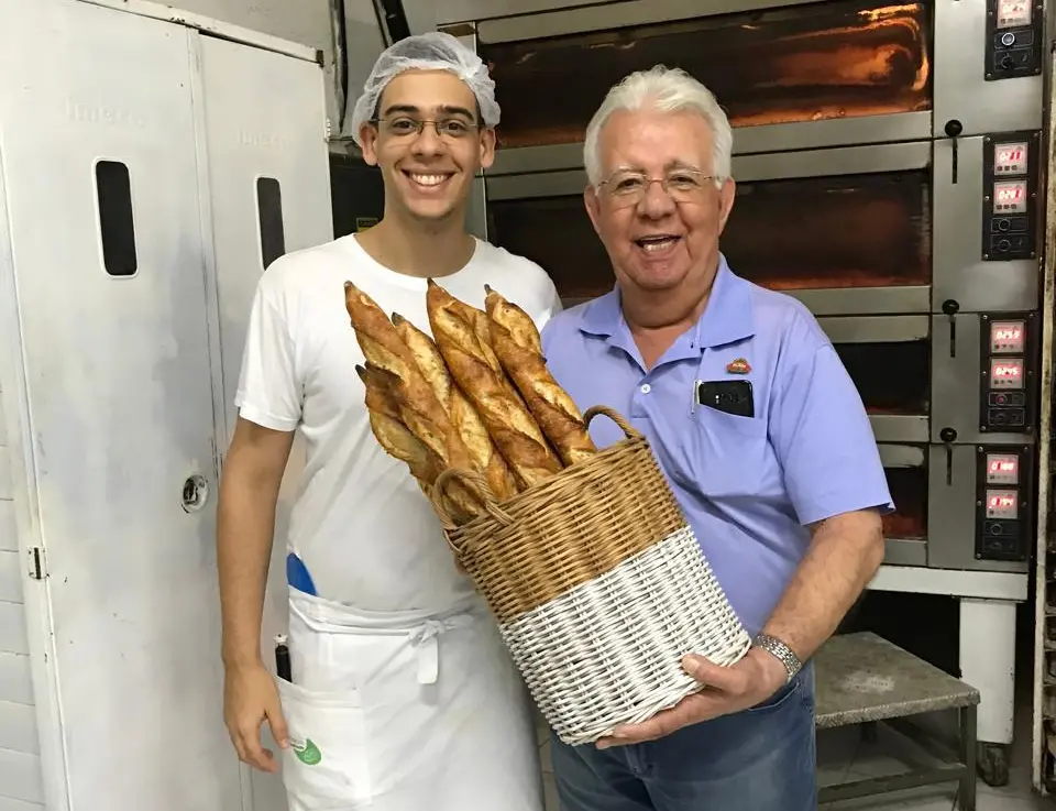 Dois homens, Brunno Malheiros e o avô, Carlos Malheiros, segurando uma cesta de baguetes em uma padaria artesanal, sorrindo para a câmera.
