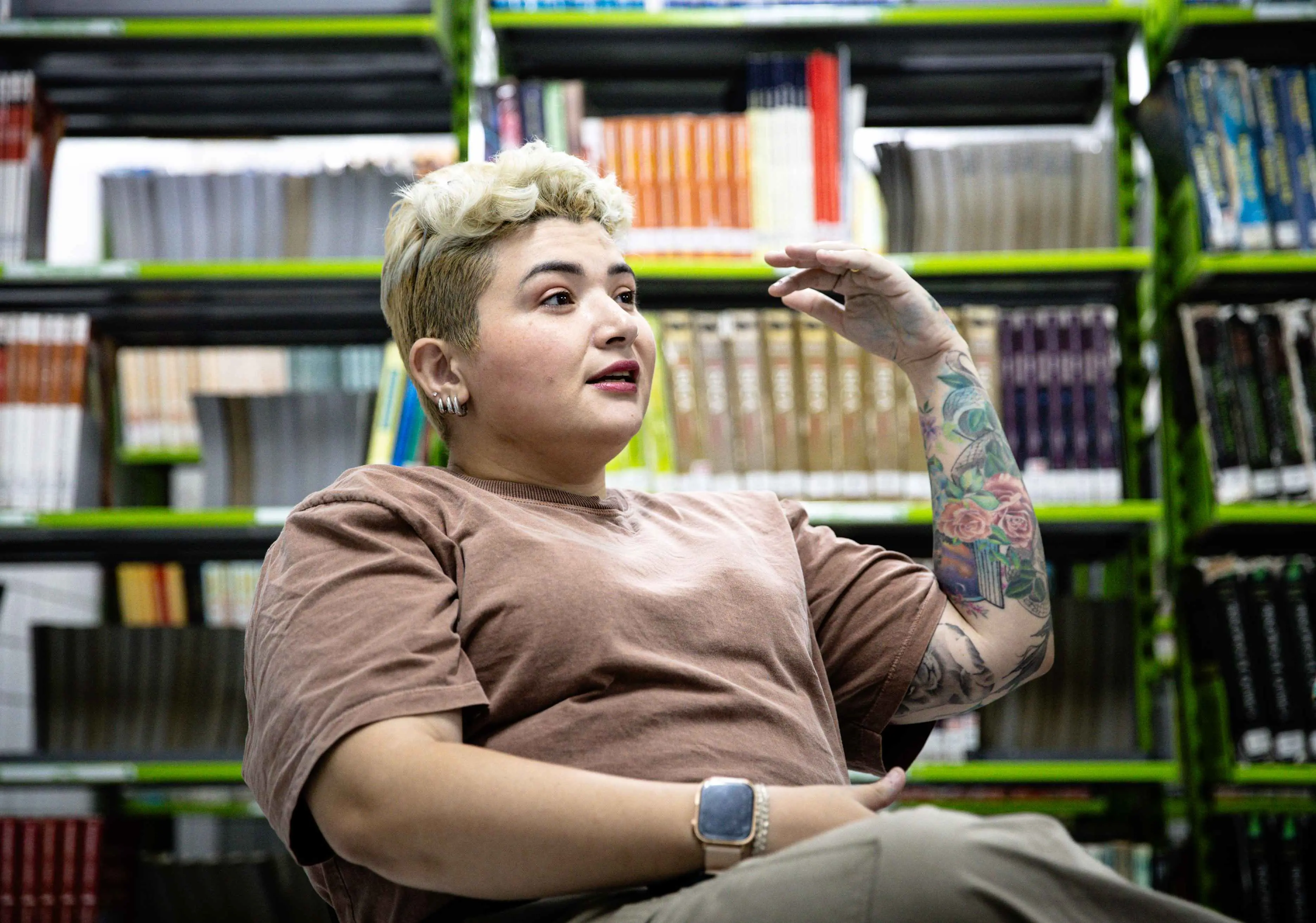 A fotografia mostra uma mulher de cabelos curtos loiros e tatuagens no braço esquerdo, sentada e gesticulando com a mão direita. Ela usa uma camiseta marrom e uma calça bege. Ao fundo, estantes de livros cheias. Imagem usada para matéria sobre cearense superdotado estar na lista de crianças prodígio