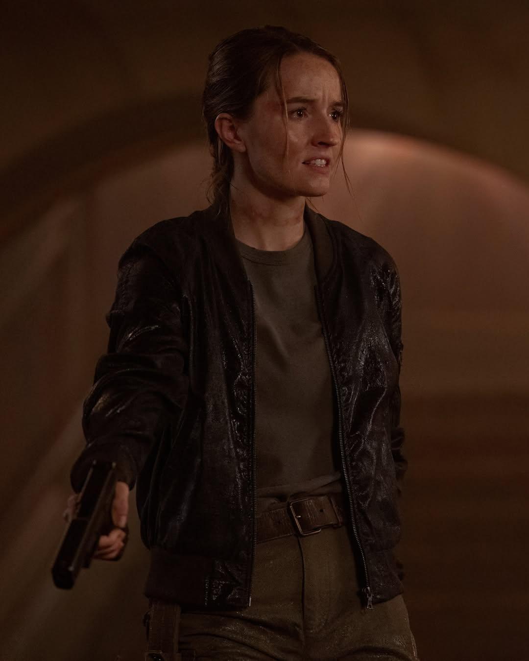 Personagem Abby, de The Last of Us, a frente de um fundo alaranjado escuro segurando uma arma e com expressão determinada para matéria que explica se Ellie morre