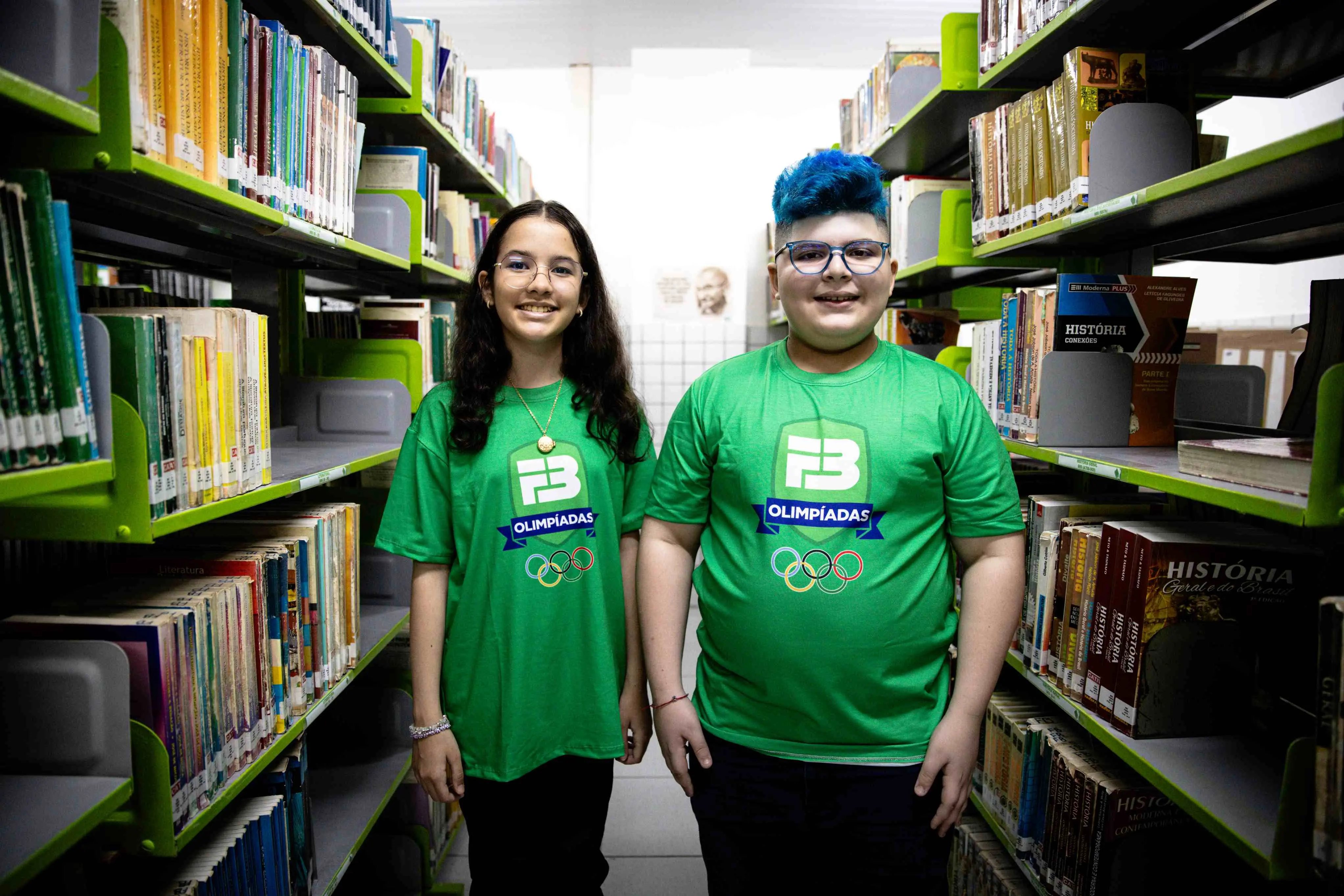 A fotografia mostra Nicole e João Pedro, dois jovens estudantes, a posarem numa biblioteca. Nicole, à esquerda, tem cabelo comprido e escuro, usa óculos e veste uma t-shirt verde com o símbolo do Farias Brito. João Pedro, à direita, tem cabelo azul vibrante, usa óculos e também veste uma t-shirt verde com o mesmo símbolo. Eles estão entre estantes de livros, com um sorriso no rosto.