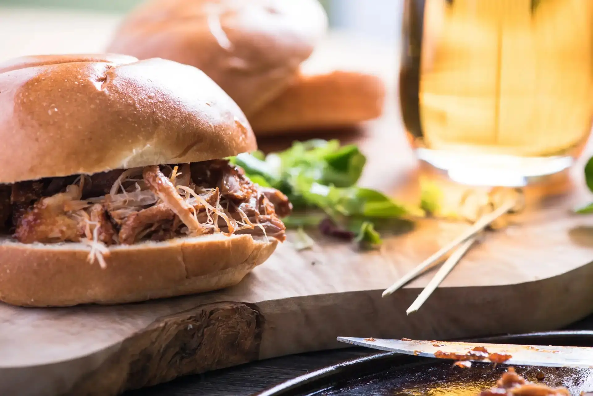 Sanduíche de carne desfiada em pão macio, acompanhado de molho, alface e uma bebida no fundo, perfeito para almoço ou lanche.