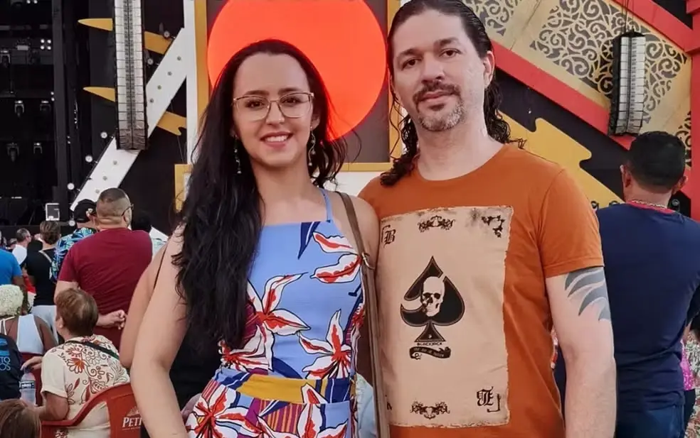 Jovem mulher e homem sorrindo em evento, com fundo decorado, pessoas ao fundo, fotografados para uma foto social, destacando-se na imagem por sua presença