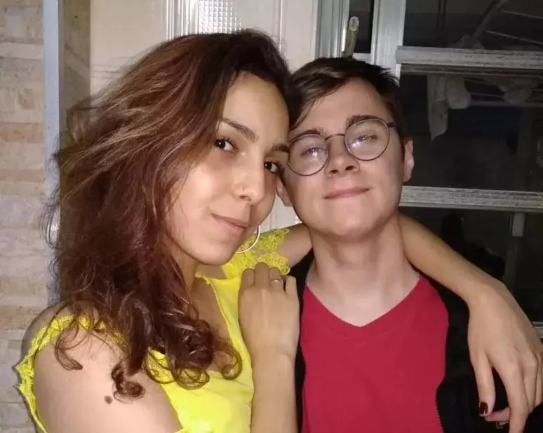 Mulher com cabelo cacheado e vestido amarelo ao lado de rapaz com óculos e camisa vermelha, ambos sorrindo e abraçados.