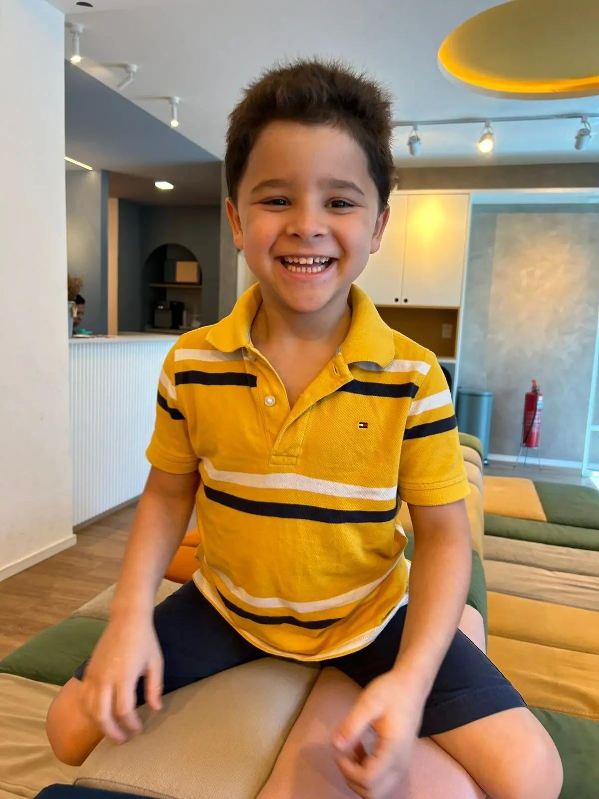 A foto mostra uma criança sentada em um banco acolchoado em um ambiente interno. Ele veste uma camisa polo listrada de amarelo e branco e um short escuro. Ao fundo, há uma parede branca, um balcão e armários.